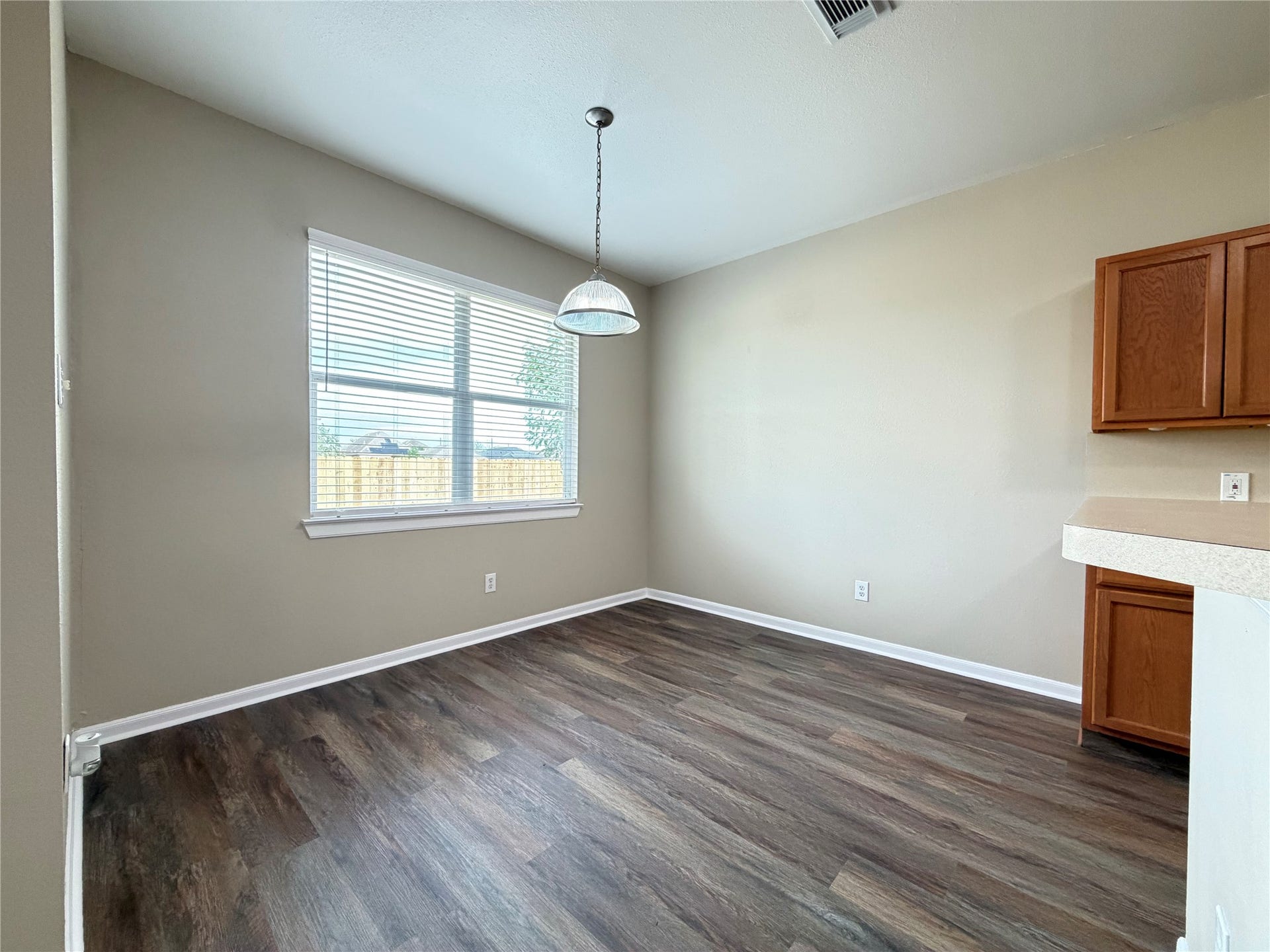 Bright dining area with large window, modern light fixture, and sleek wood flooring, adjacent to kitchen.