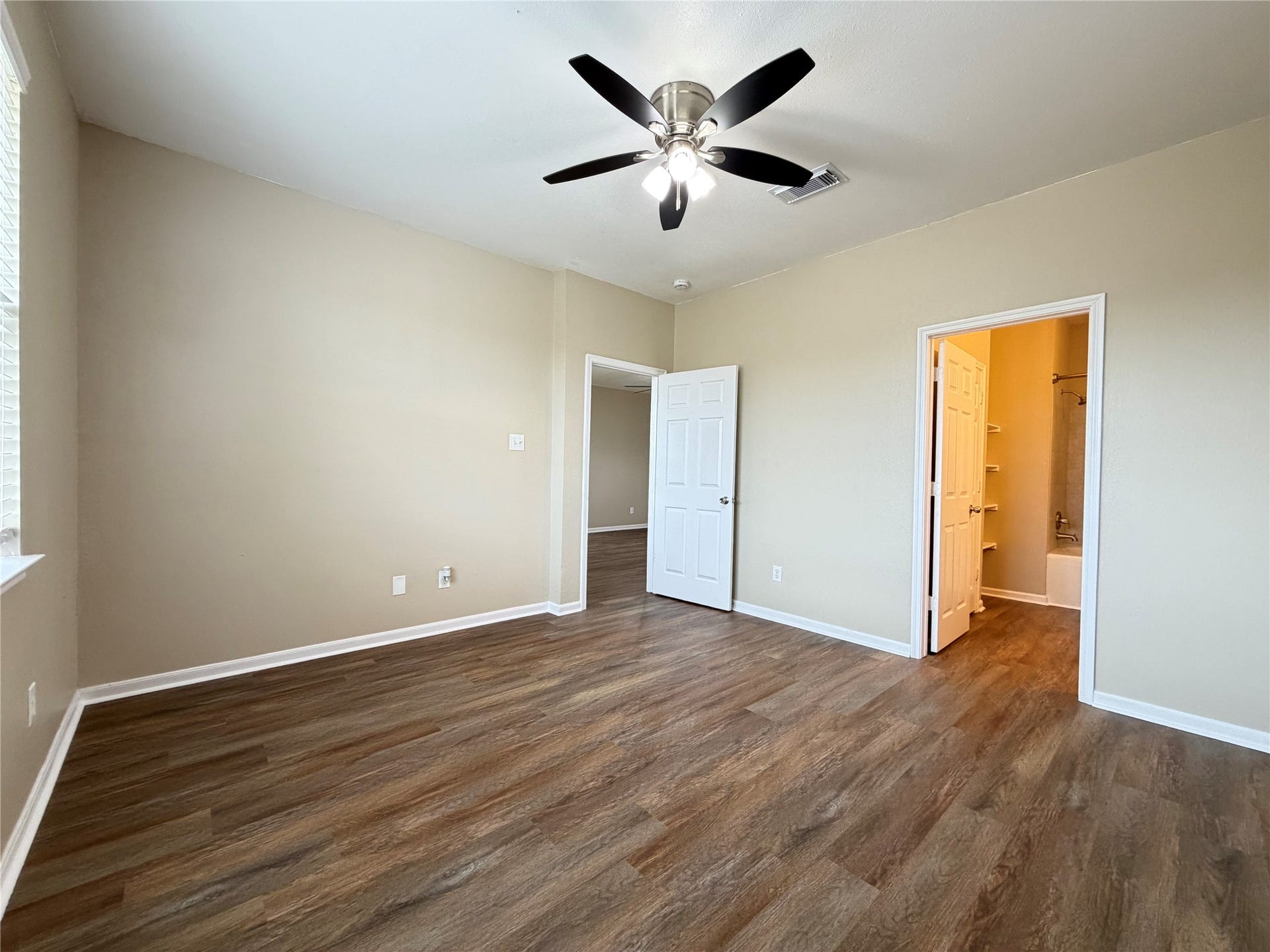 Spacious primary bedroom with warm wood flooring, neutral walls, and modern ceiling fan, leading to an en-suite bathroom for added convenience.