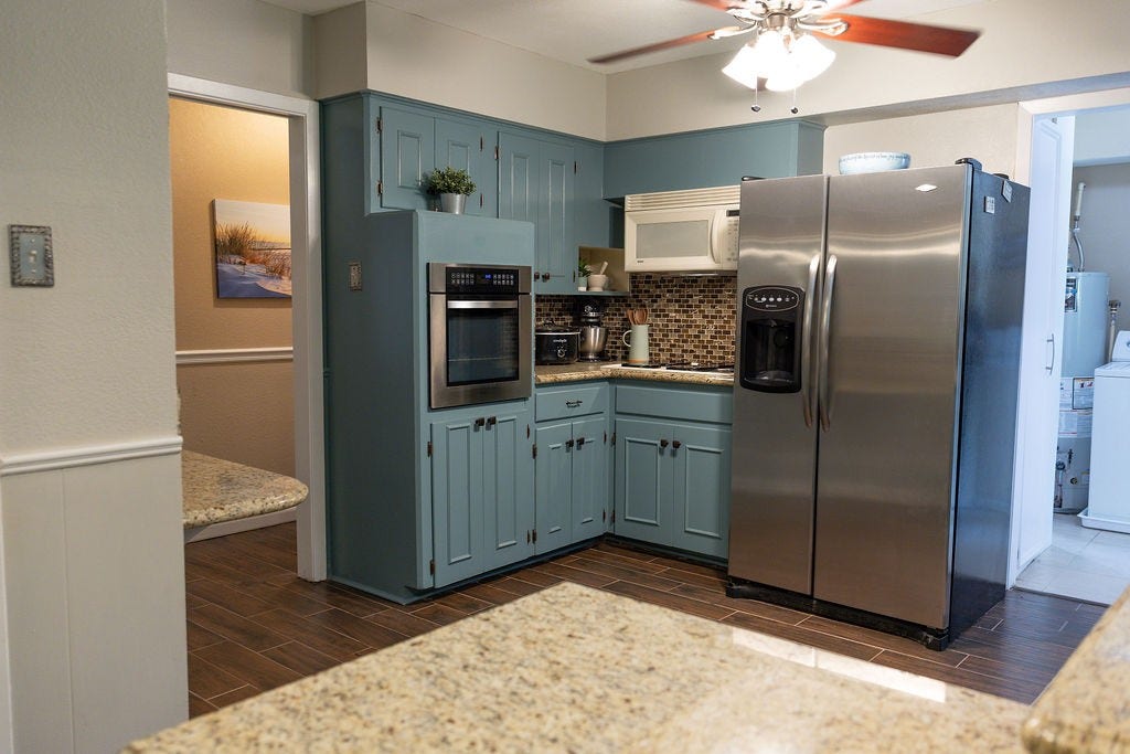 1314 Laurel Street, La Marque, TX 77568 This kitchen features stylish blue cabinets, stainless steel appliances, and granite countertops. The mosaic tile backsplash adds a modern touch, and the ceiling fan provides comfort. Adjacent areas offer additional space and functionality.