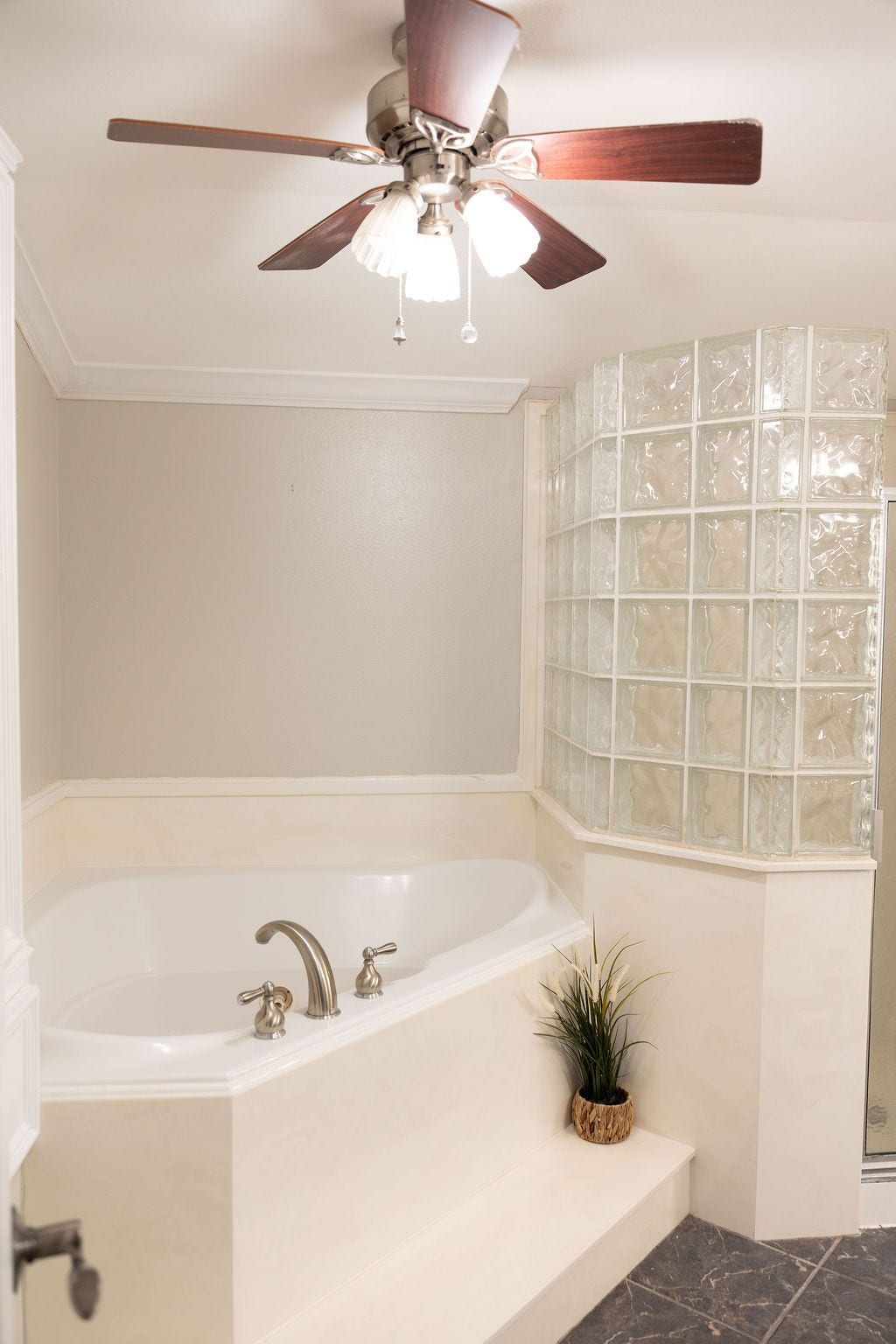 1314 Laurel Street, La Marque, TX 77568 This bathroom features a spacious corner bathtub with modern fixtures, surrounded by elegant glass block walls. A ceiling fan with lights adds comfort, while a small plant provides a touch of greenery.