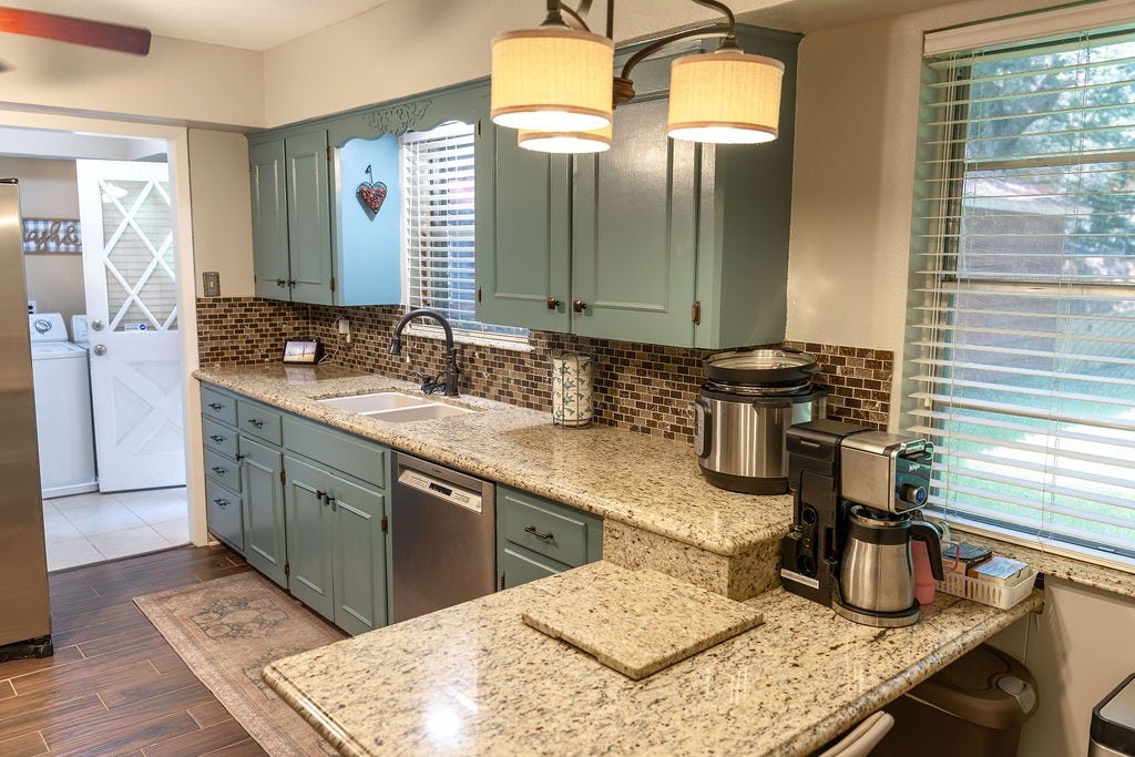 1314 Laurel Street, La Marque, TX 77568 This kitchen features stylish light blue cabinets, granite countertops, and a mosaic tile backsplash. It's equipped with modern appliances and offers ample natural light through a large window. There's a spacious connection to the laundry area.