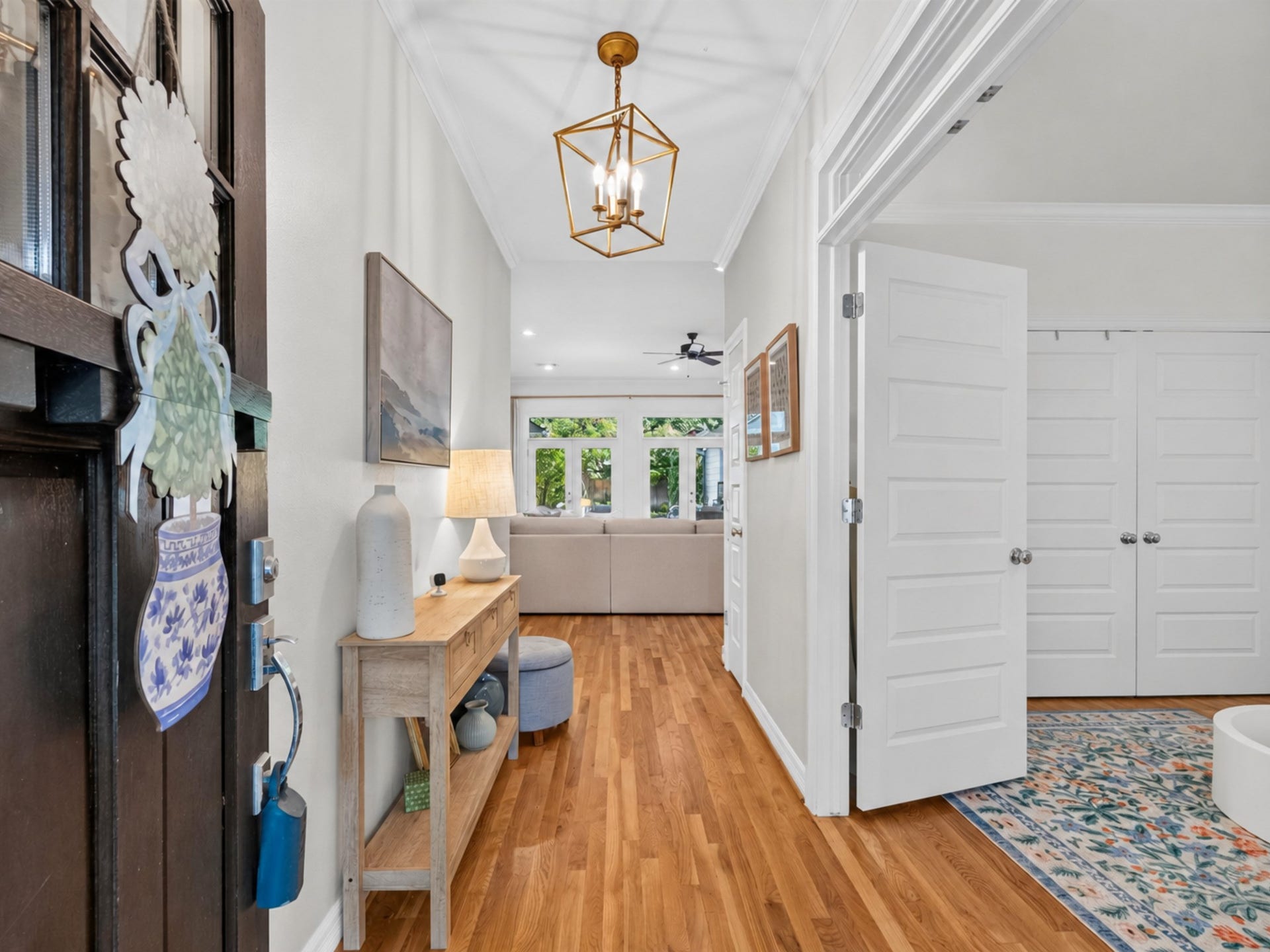 An inviting entryway with warm wood floors leads to a bright living area, featuring stylish lighting and a cozy living space. A side room offers versatility and charm.