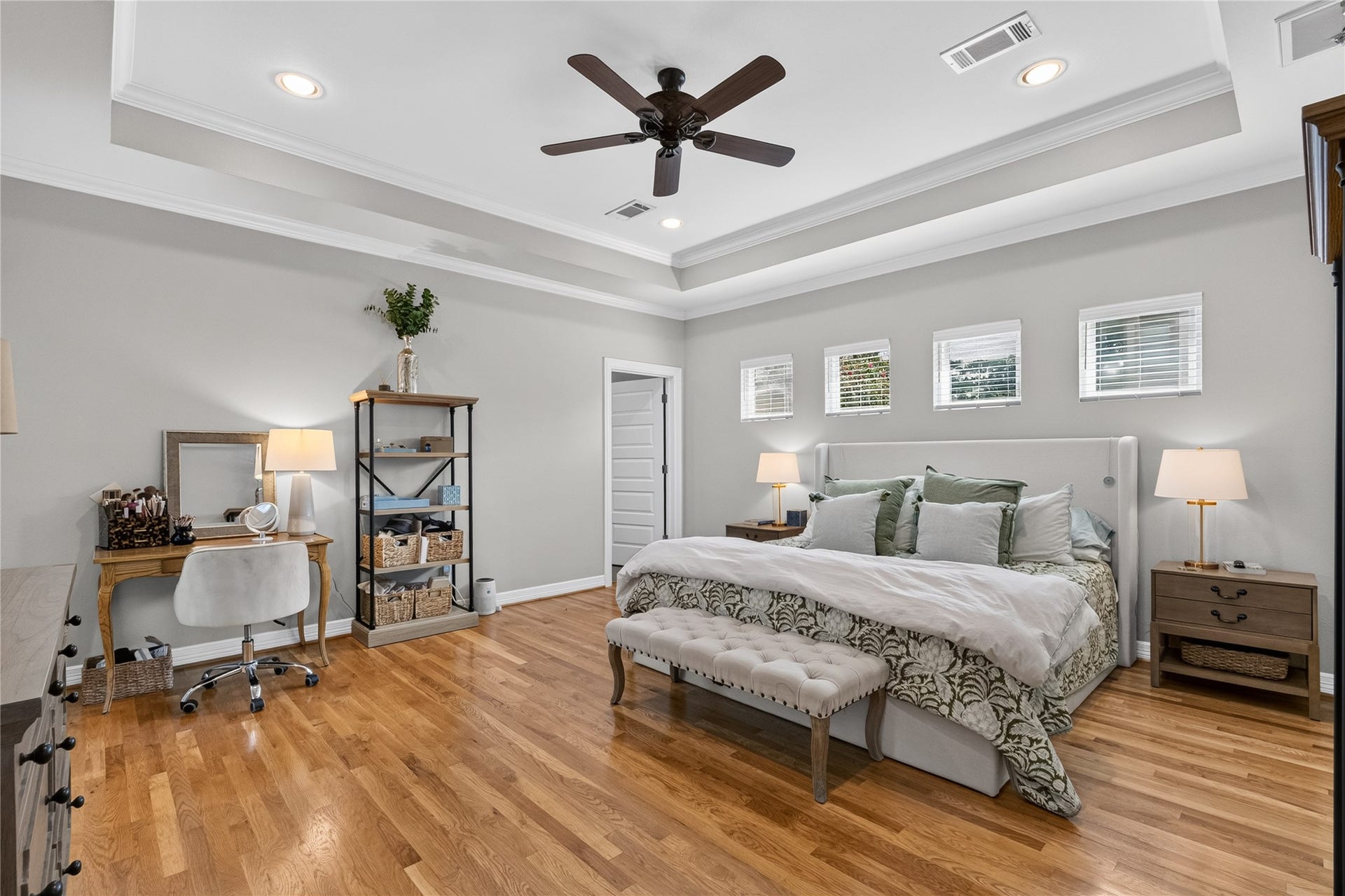 Spacious and elegant primary bedroom featuring hardwood floors, a tray ceiling with recessed lighting, and a ceiling fan.  Natural light streams through multiple windows, creating a bright and inviting atmosphere.
