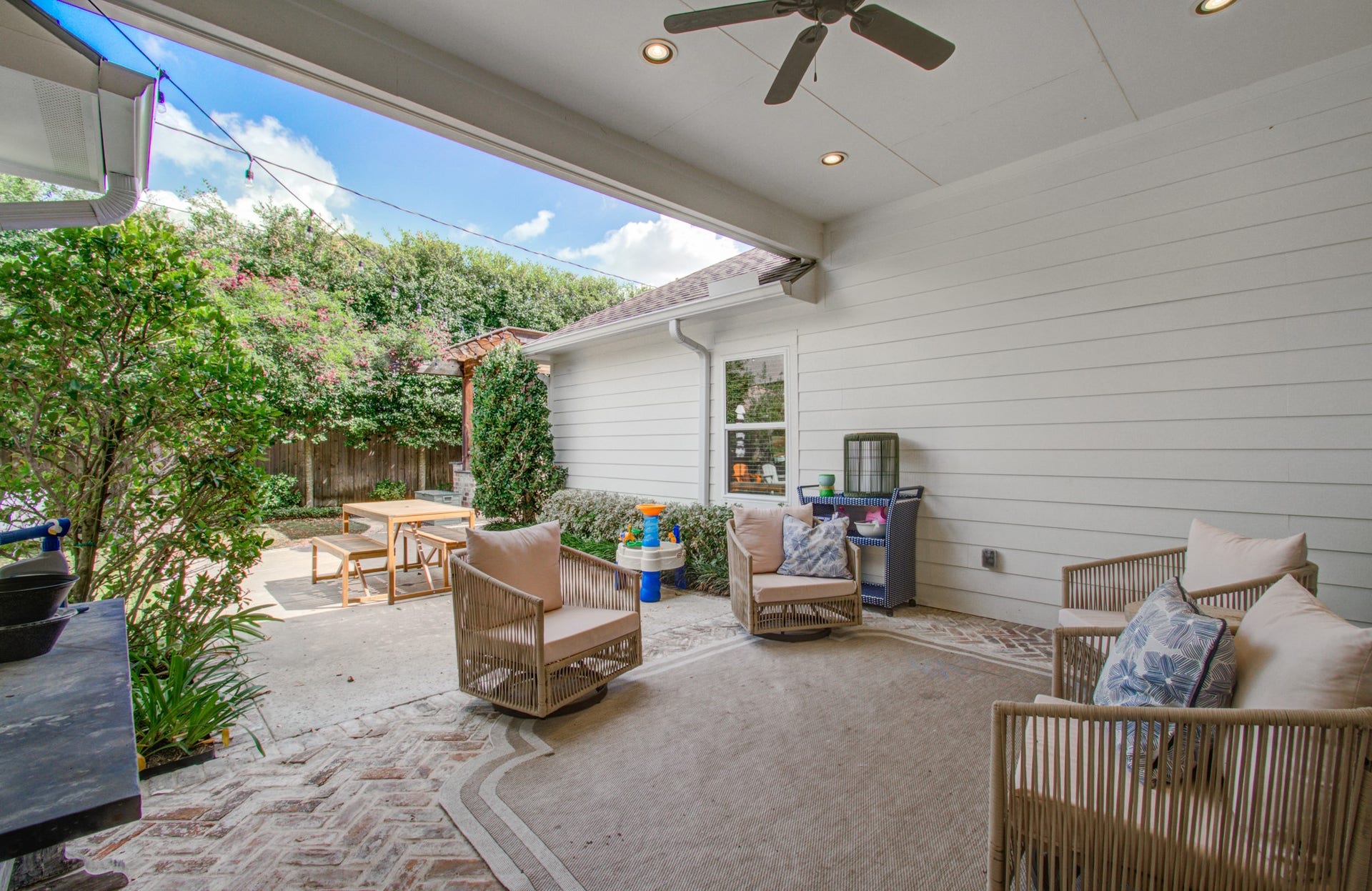 Charming covered outdoor patio with comfortable seating, ceiling fan, and lush greenery, perfect for relaxing or entertaining.