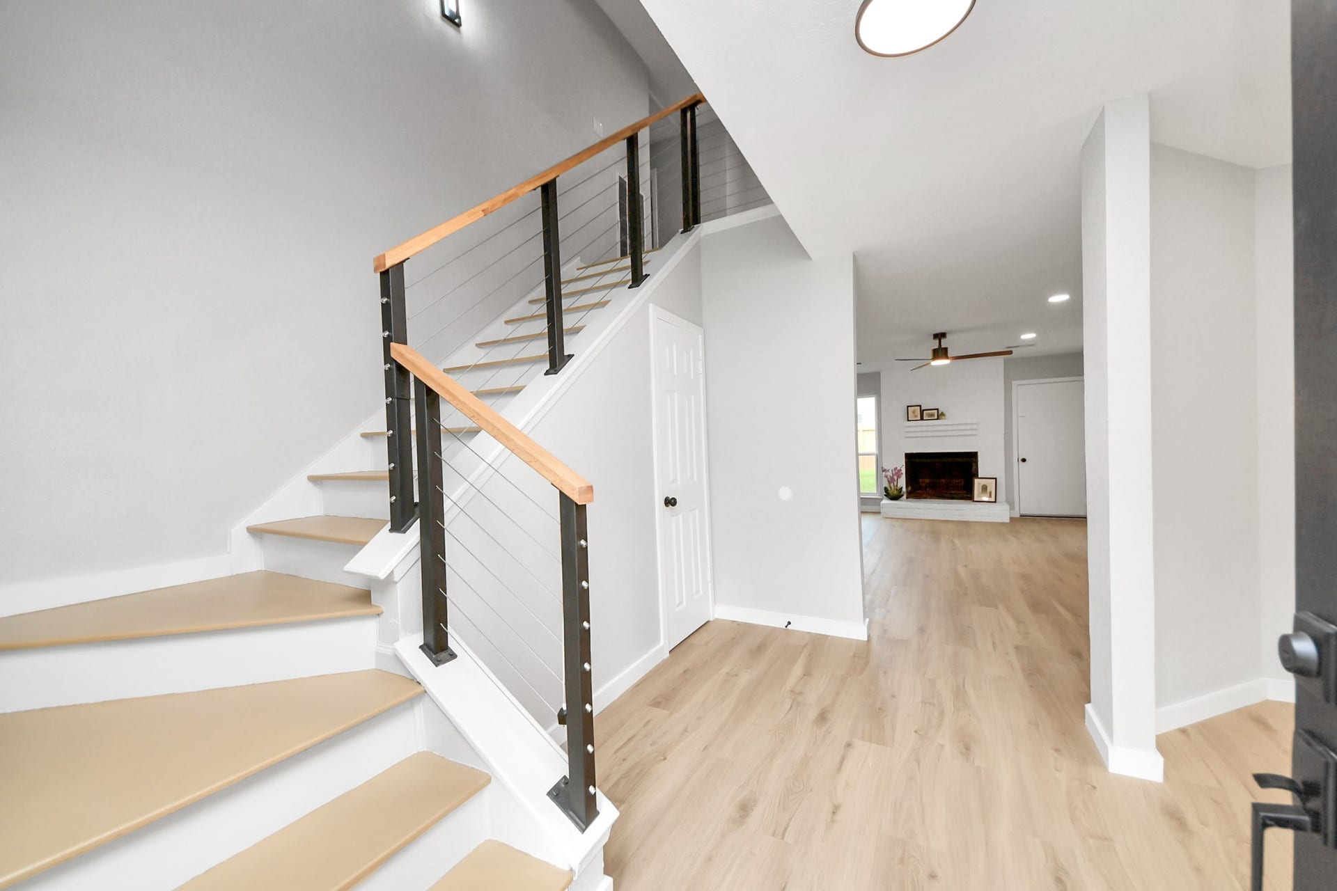 3234 Pebble Lake Drive, Sugar Land, TX 77479 This photo showcases a bright, modern entryway with sleek wood and metal staircase railing, light wood flooring, and an open view into a cozy living room with a fireplace.