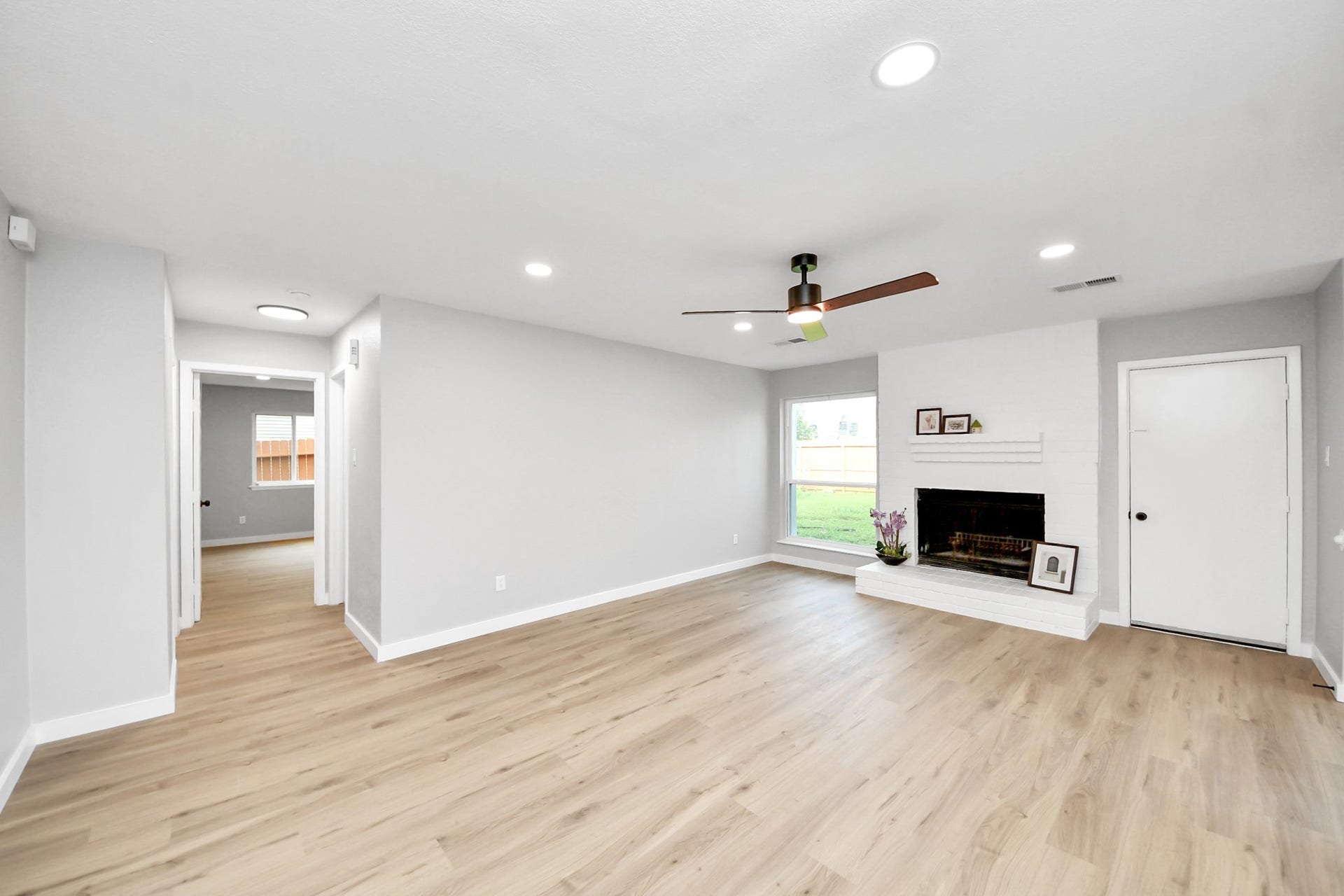 3234 Pebble Lake Drive, Sugar Land, TX 77479 This spacious living room features light wood flooring, a modern ceiling fan, and a cozy fireplace framed by a white brick mantel. The room is well-lit with recessed lighting and a large window providing natural light. A hallway leads to additional rooms, offering an open and inviting layout.