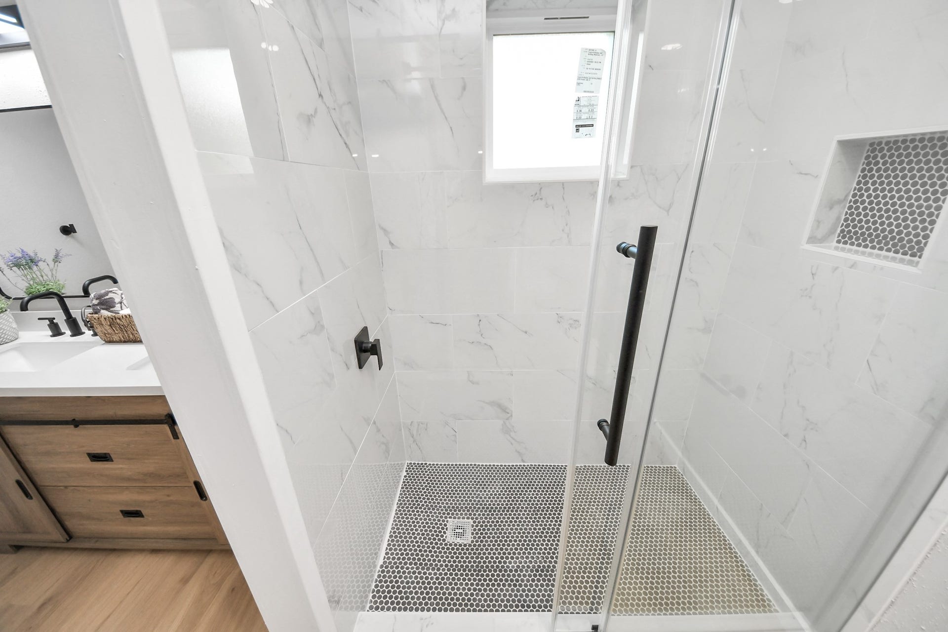 3234 Pebble Lake Drive, Sugar Land, TX 77479 Modern bathroom with a glass-enclosed shower featuring marble-look tiles and a niche. There's a sleek black handle and fixtures, a small window for natural light, and a glimpse of a wooden vanity with a white countertop.