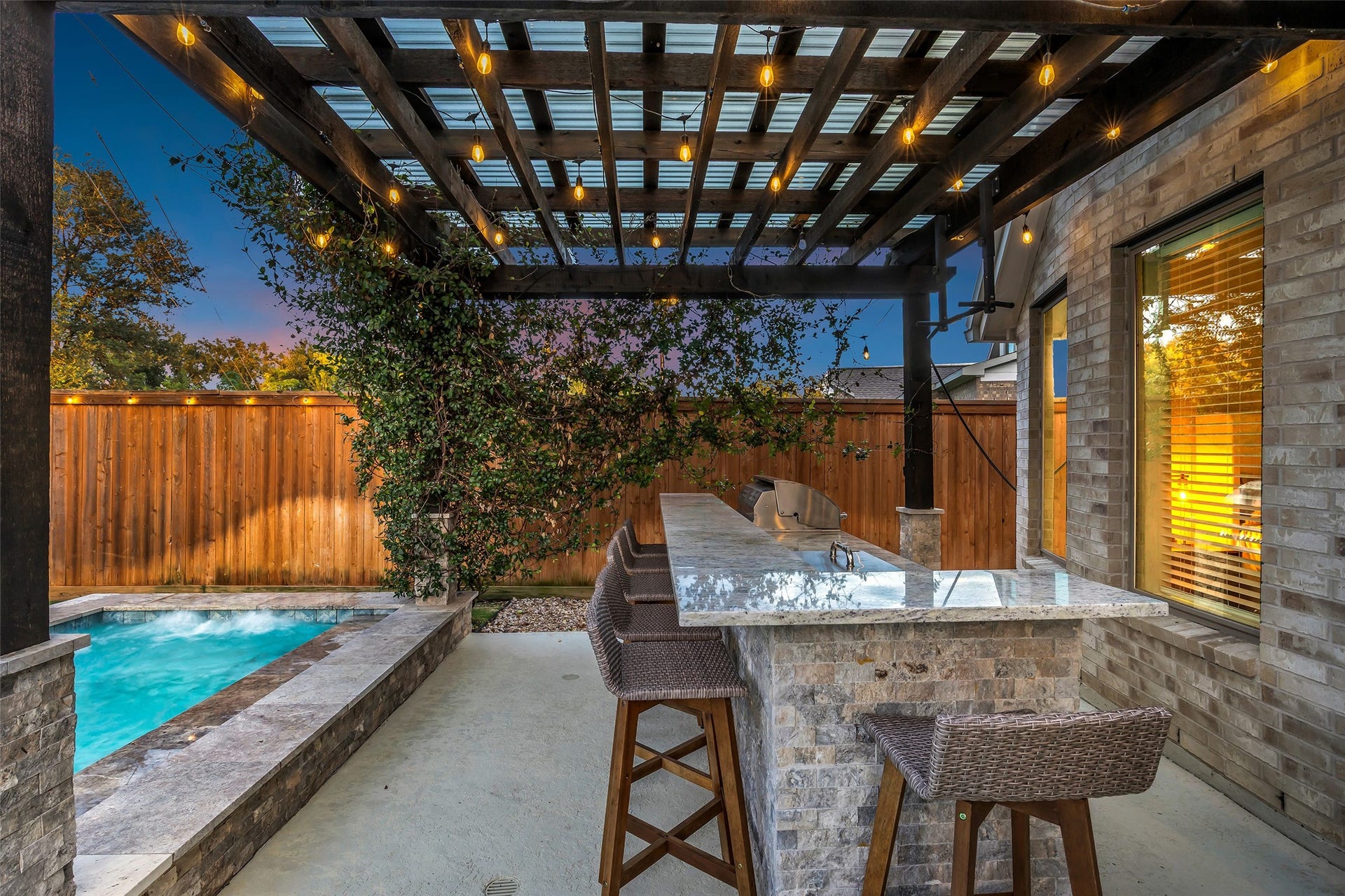 Gather at the stone outdoor bar under a string-lit pergola, overlooking the spa and pool.