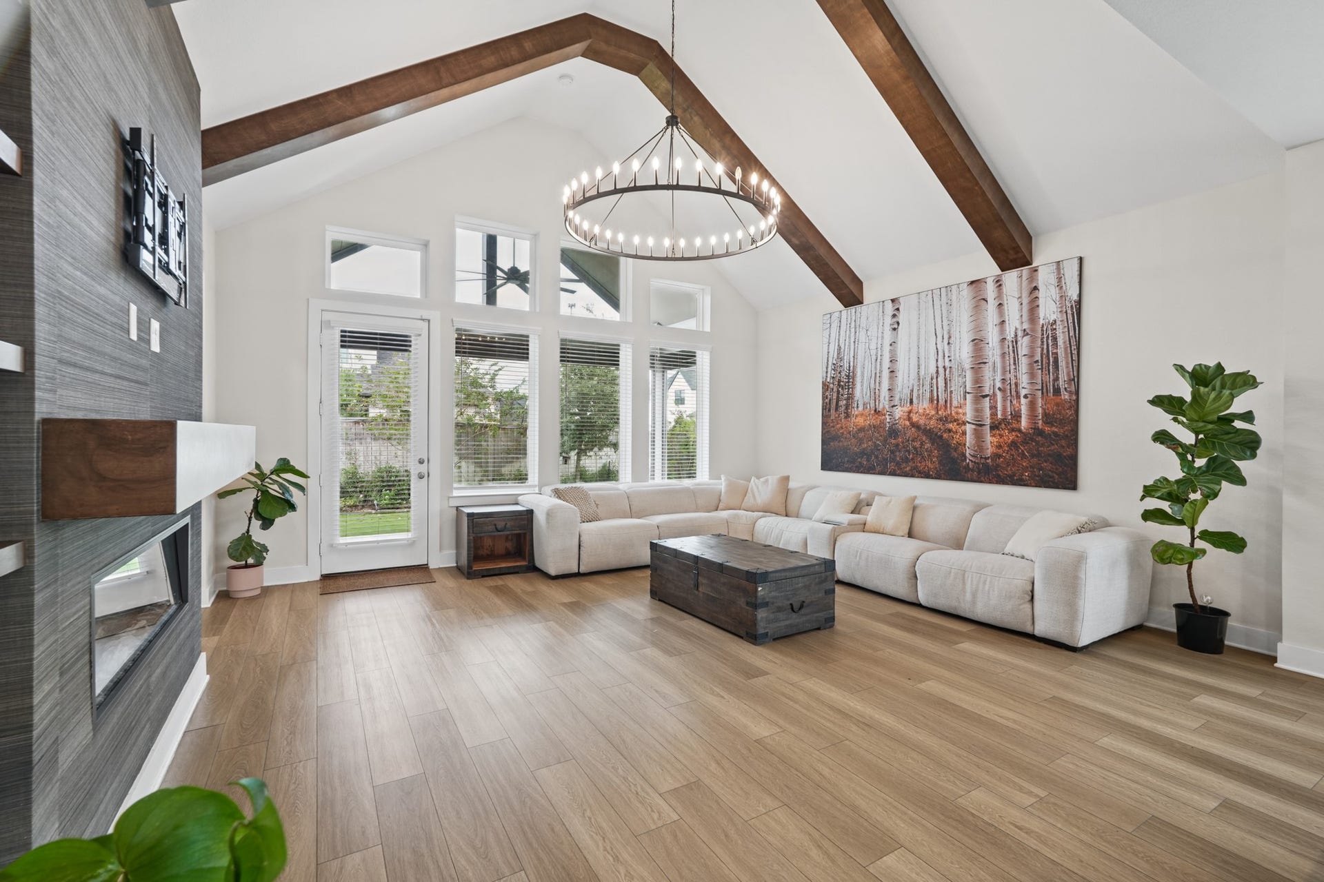 The living room centers on a fireplace and built-in media wall beneath vaulted ceilings and wood beams, opening to the kitchen on one side and the covered patio on the other.