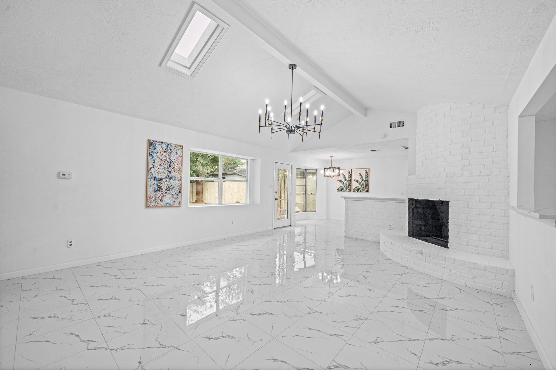 12123 Pompano Lane, Houston, TX 77072 Bright and spacious living area featuring a vaulted ceiling with skylight, elegant chandelier, and a cozy white brick fireplace, complemented by glossy marble-style flooring and large windows for abundant natural light.