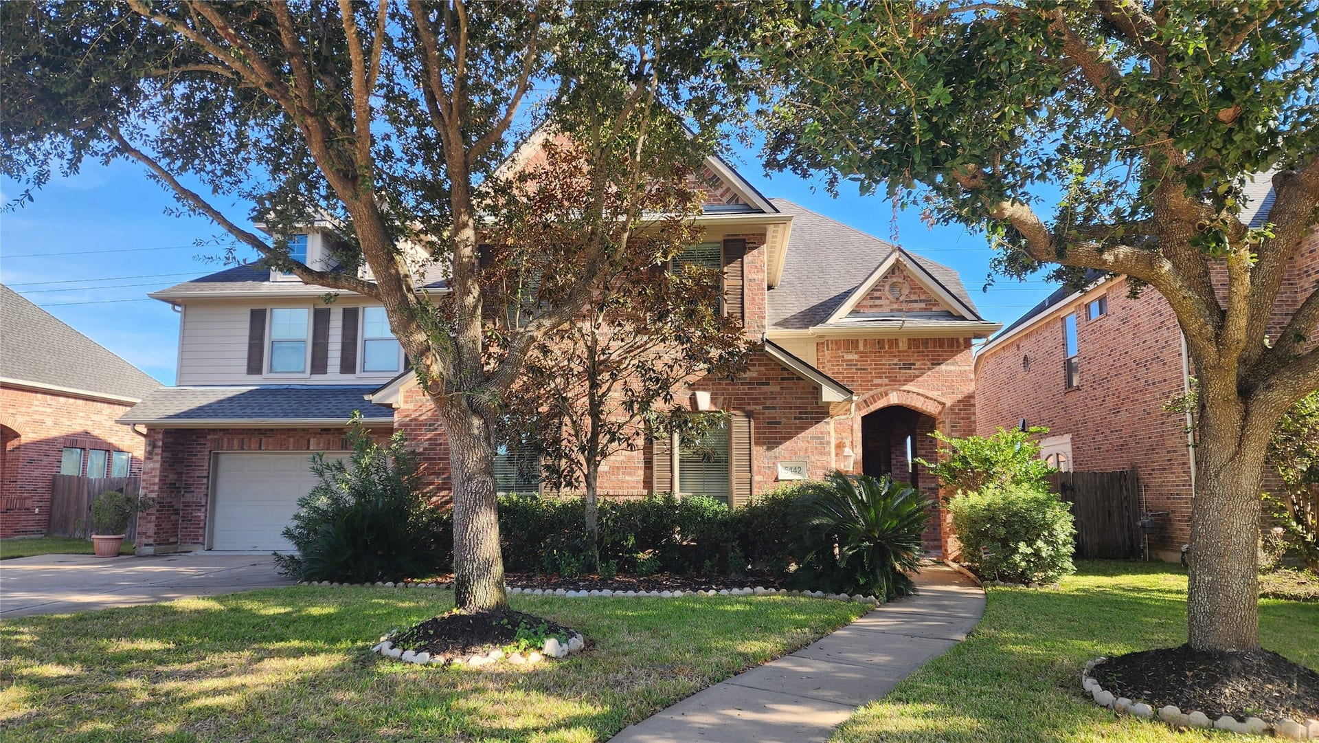 5442 Faircreek Ln Lane, Katy, TX 77450 Charming two-story brick home with a welcoming front entrance, nestled among mature trees and lush landscaping, featuring a spacious driveway leading to a two-car garage.