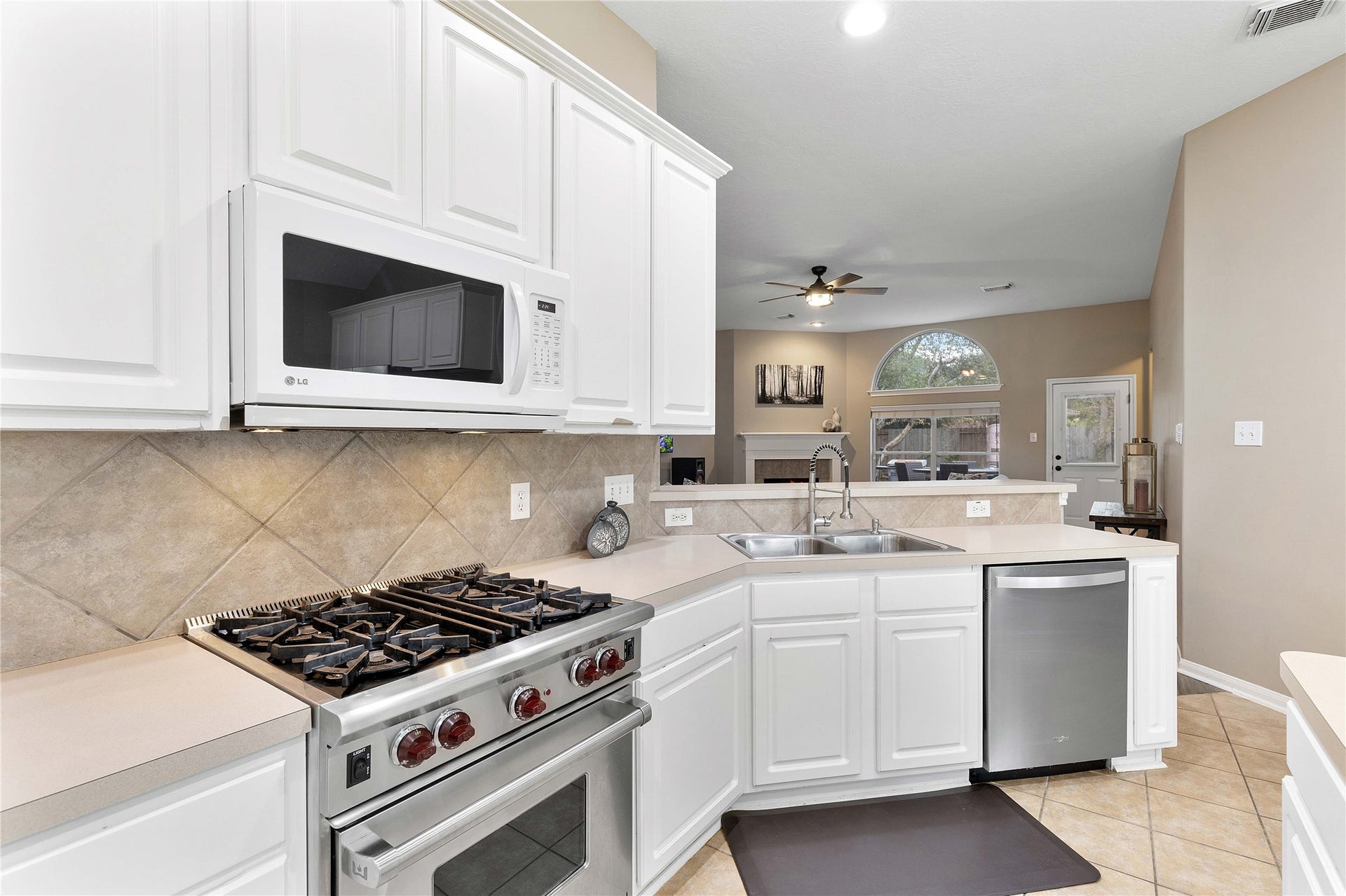 18343 Otter Creek Trail, Humble, TX 77346 Charming kitchen with white cabinetry, stainless steel appliances, and a spacious countertop overlooking a cozy living area with a fireplace and large arched window.