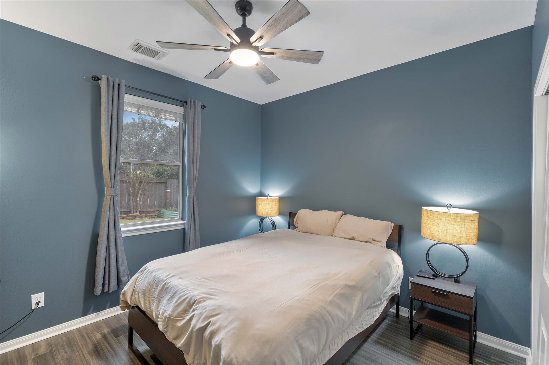 18343 Otter Creek Trail, Humble, TX 77346 Cozy bedroom with calming blue walls, featuring a modern ceiling fan, sleek flooring, and ample natural light from the window overlooking the backyard.