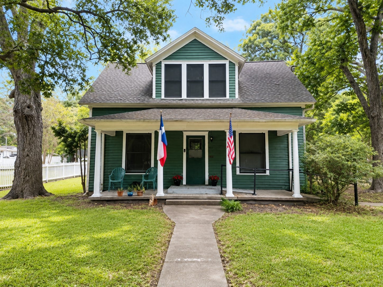 Charming green exterior with crisp white trim, a welcoming covered front porch, and a mature tree canopy that adds natural character to the front yard. A paved walkway provides a direct approach to the entry.