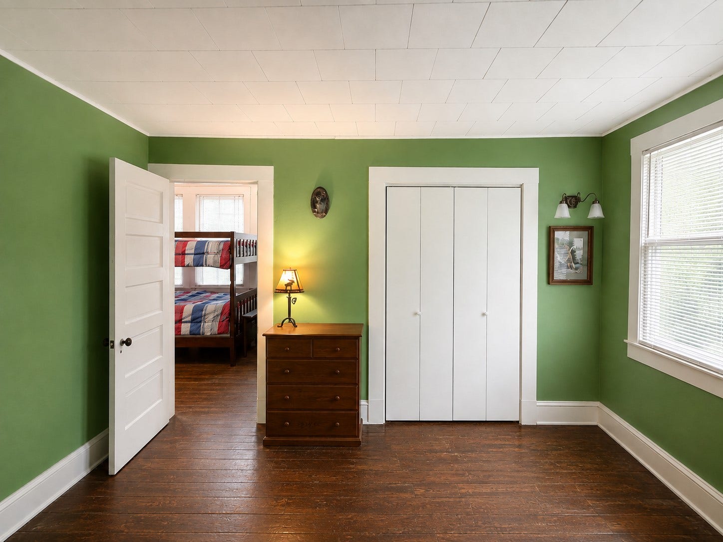 Spacious upstairs bedroom features rich wood flooring, green walls, classic white trim, and a large closet. A wide window brings natural light into the room, while the open floor area allows for flexible furniture placement.