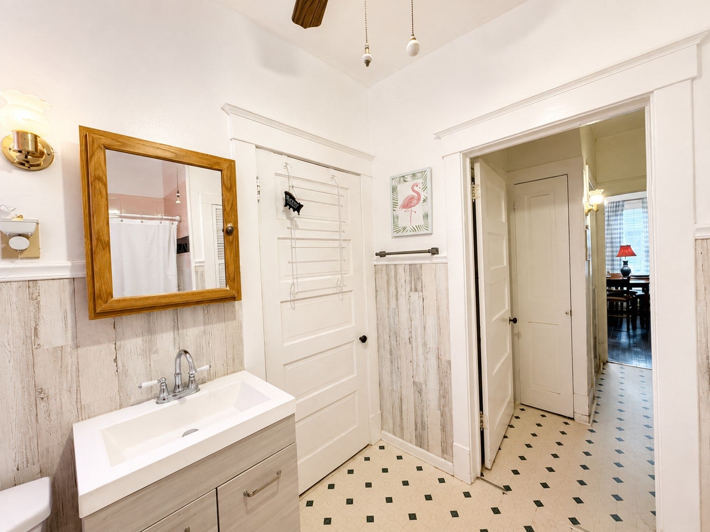 This angle highlights the updated vanity, framed mirror, classic paneled doors, and distinctive wood-look wall treatment. The adjoining doorway provides a clear view of the bathrooms connection to the surrounding living spaces.