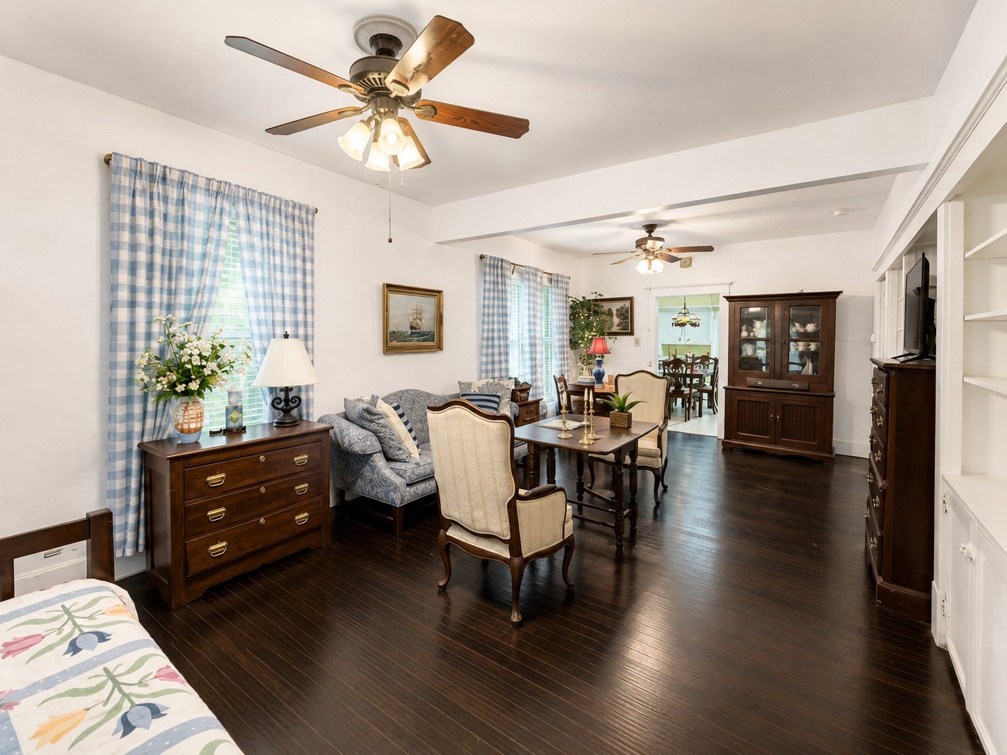 An elongated living space offers flexible room for multiple seating or functional areas, with several windows bringing in natural light. Dark wood flooring, a ceiling fan, and classic trim carry a cohesive look throughout.