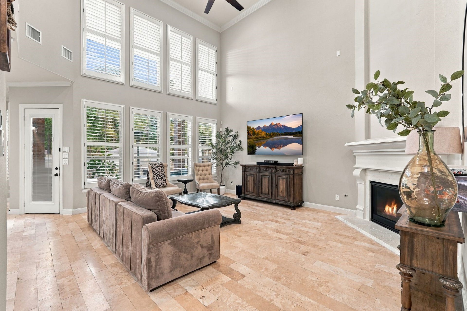 13714 Arcott Bend Drive, Tomball, TX 77377 Breathtaking two-story living room showcases soaring ceilings, a dramatic wall of windows adorned with plantation shutters, an elegant fireplace, and beautiful travertine flooring. An abundance of natural light enhances the home's sophisticated architectural design, creating a stunning space for luxurious everyday living and entertaining.
