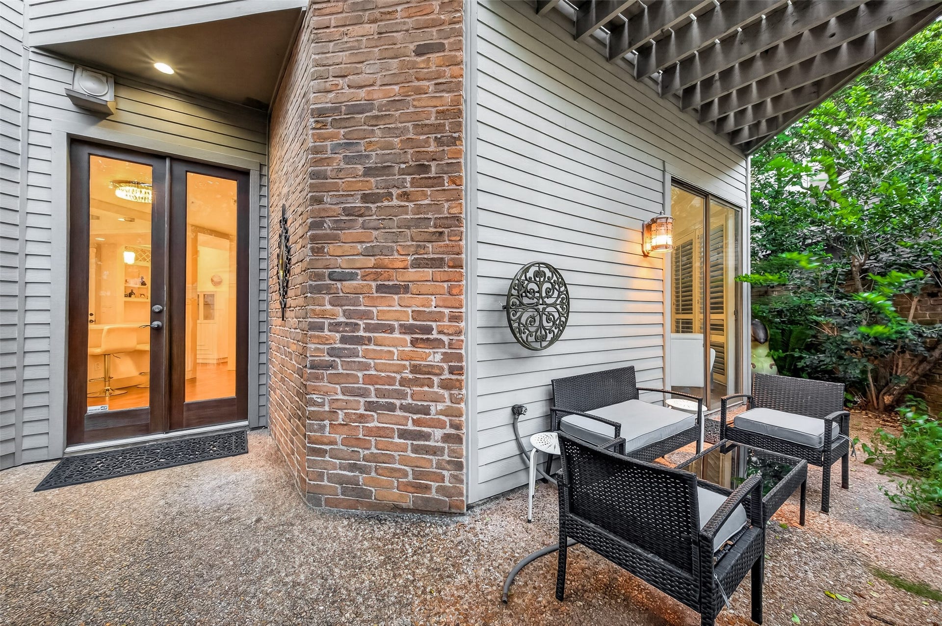 This photo showcases a cozy outdoor patio with modern wicker furniture and a glass-top table, set against a backdrop of brick and siding. A glass door provides entry to the interior, while lush greenery surrounds the area, creating a private and inviting space.