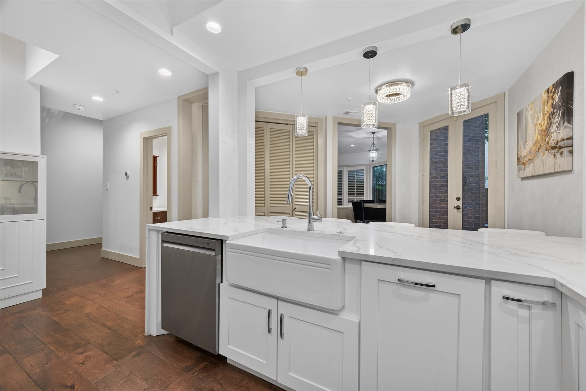 This modern kitchen features sleek white cabinetry, a large farmhouse sink, and elegant marble countertops. It boasts stainless steel appliances, stylish pendant lighting, and engineer wood  flooring, with an open view into the dining area through glass doors.