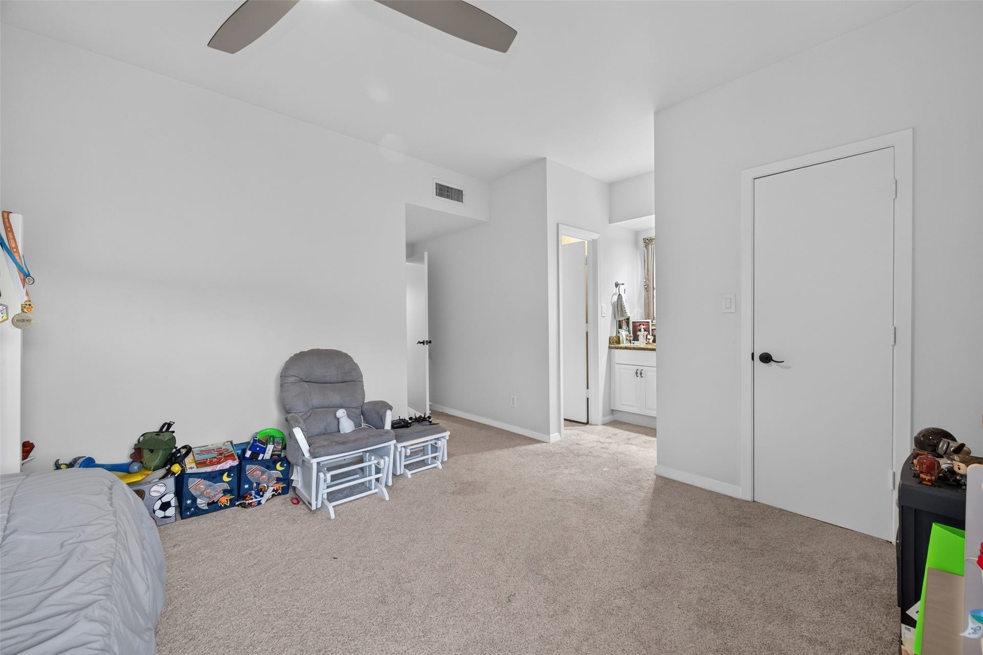 This photo shows a spacious, carpeted bedroom with neutral-colored walls. It features a ceiling fan, a cozy chair, and a glimpse of an adjoining bathroom with a vanity. The room is well-lit and has a tidy, inviting atmosphere.