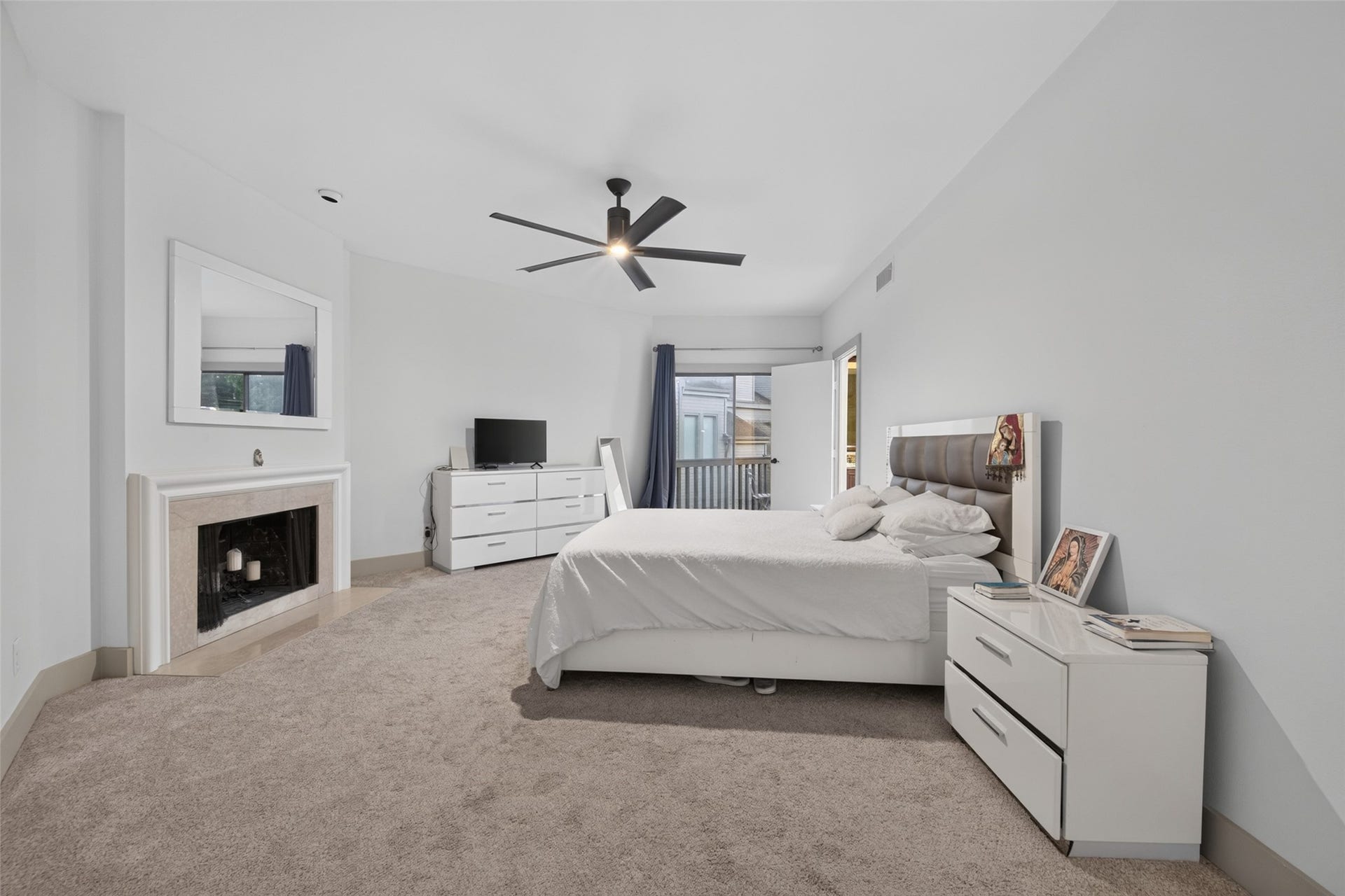 This spacious bedroom features neutral carpeting, a cozy fireplace, modern ceiling fan, and ample natural light from the sliding glass door leading to a balcony. The room is complemented by sleek white furniture, creating a clean and inviting atmosphere.
