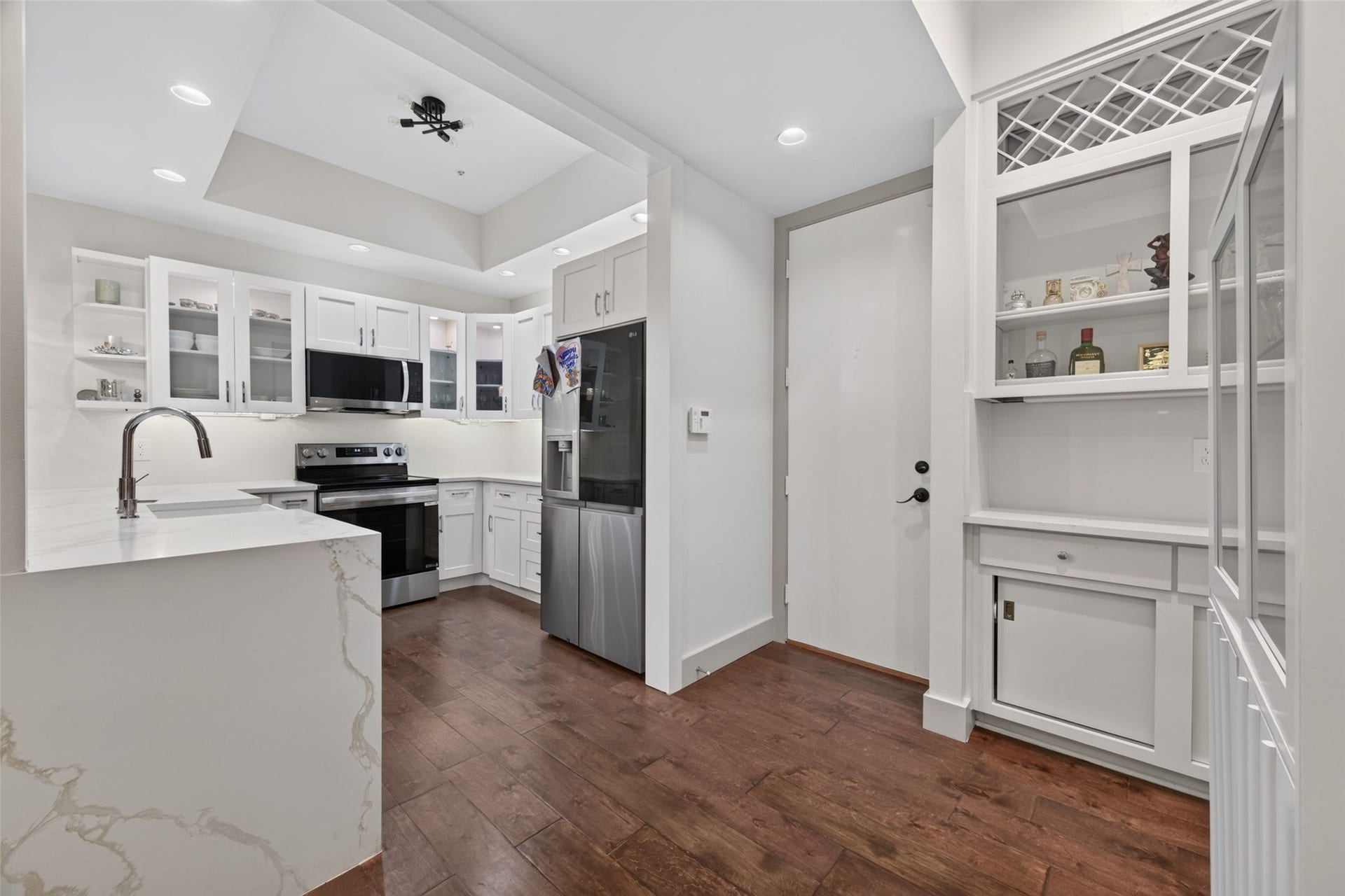 This modern kitchen features sleek white cabinetry, stainless steel appliances, and a marbled countertop with a breakfast bar. The engineer wood  flooring adds warmth, while recessed lighting and open shelving enhance the space's contemporary look.