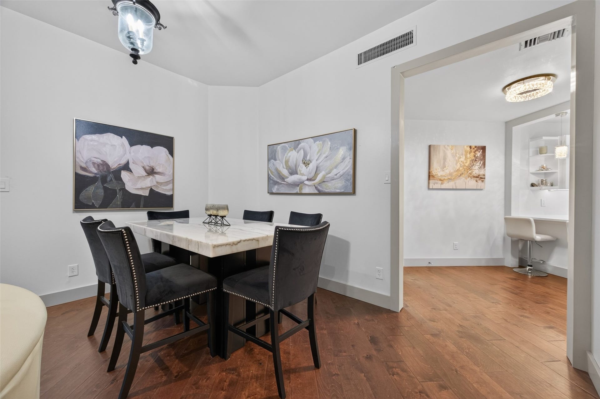 This photo showcases a stylish dining area featuring a marble table surrounded by six elegant black chairs. The room is adorned with floral artwork, has warm wood flooring, and opens into a cozy, well-lit study nook with built-in shelving.