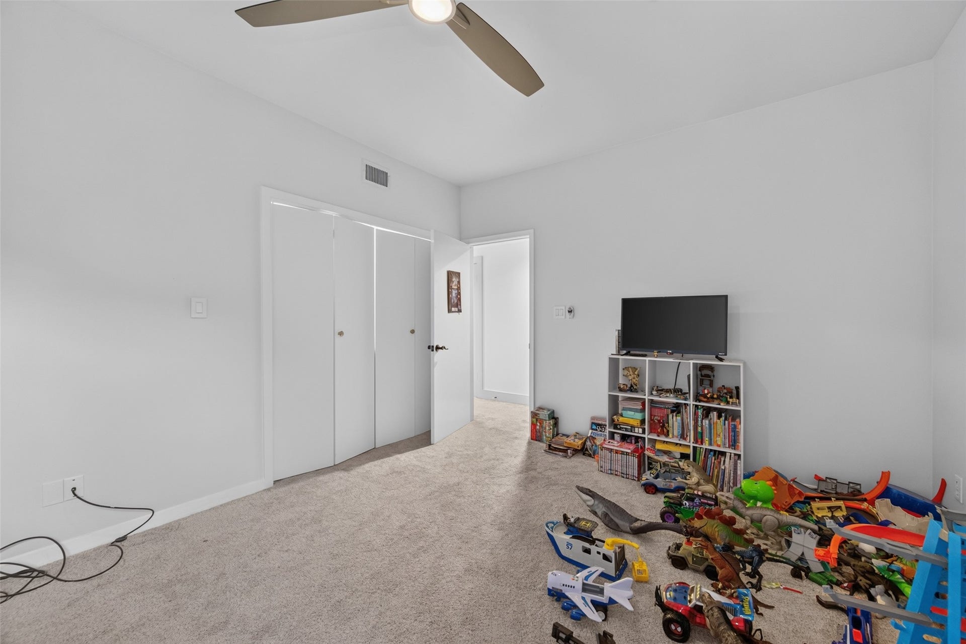 This photo shows a cozy room with carpeted flooring and neutral walls. It features a ceiling fan, a closet with sliding doors, and a small entertainment setup with a TV and shelves. There are toys and books on the shelves and floor, indicating it's used as a playroom or child's bedroom.