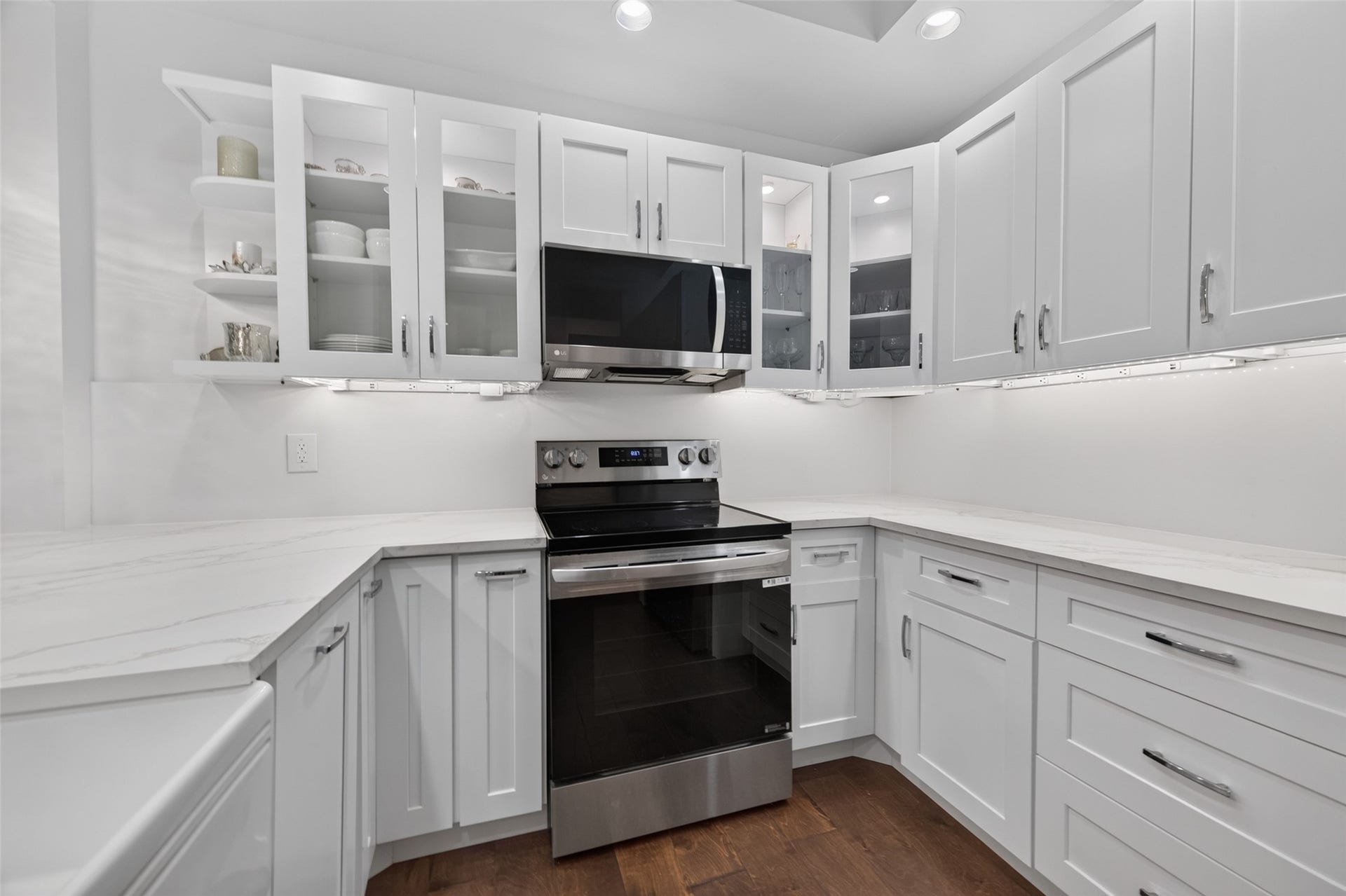 This modern kitchen features sleek white cabinetry, glass-front upper cabinets, stainless steel appliances, and under-cabinet lighting. It offers a clean, bright look with ample counter space and storage.