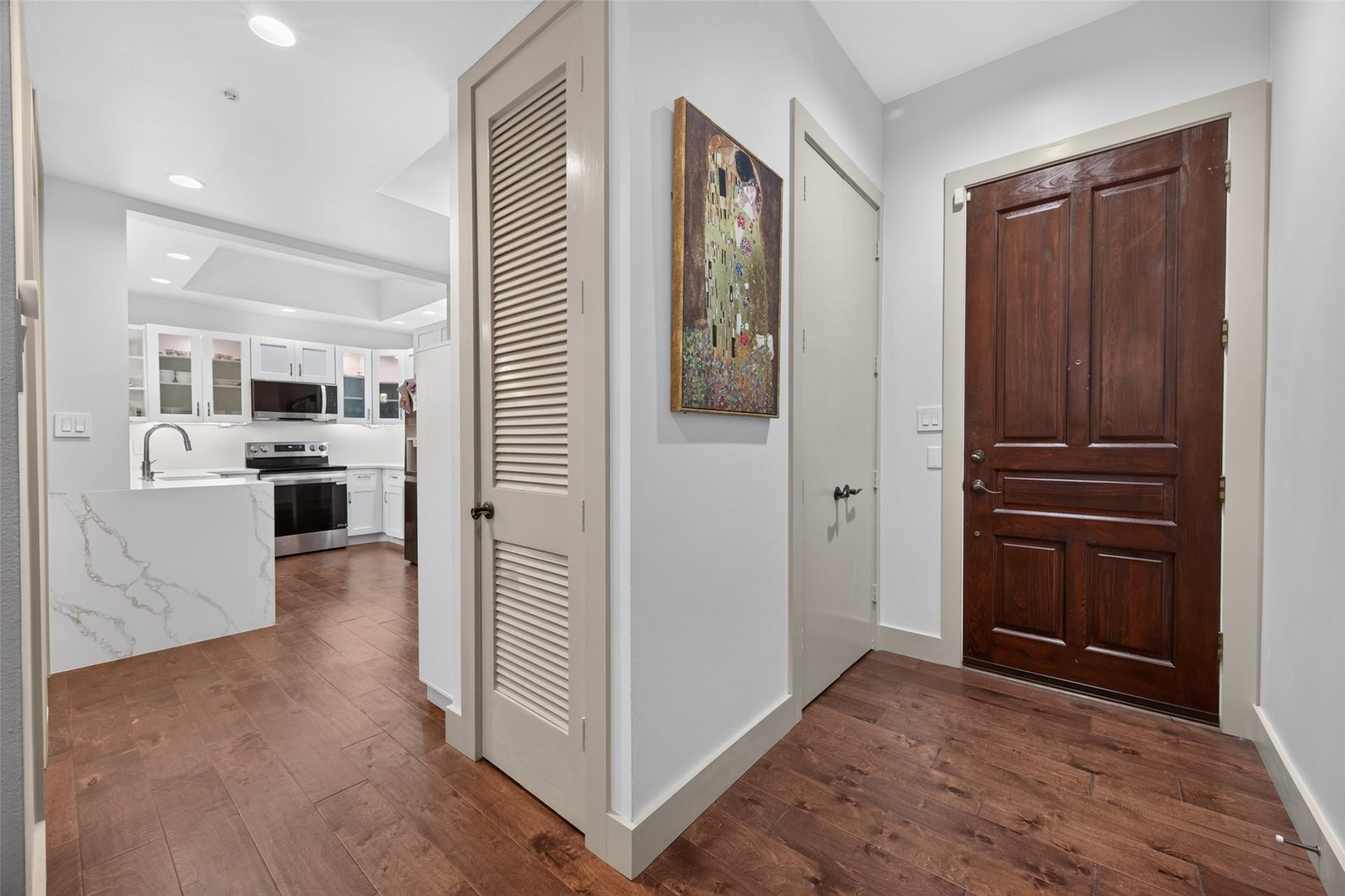 This photo showcases a modern entryway with rich engineer wood flooring and a solid wooden front door. It leads to a stylish kitchen featuring sleek white cabinetry, a marble-look countertop, and stainless steel appliances. The space is well-lit and includes a decorative wall piece.