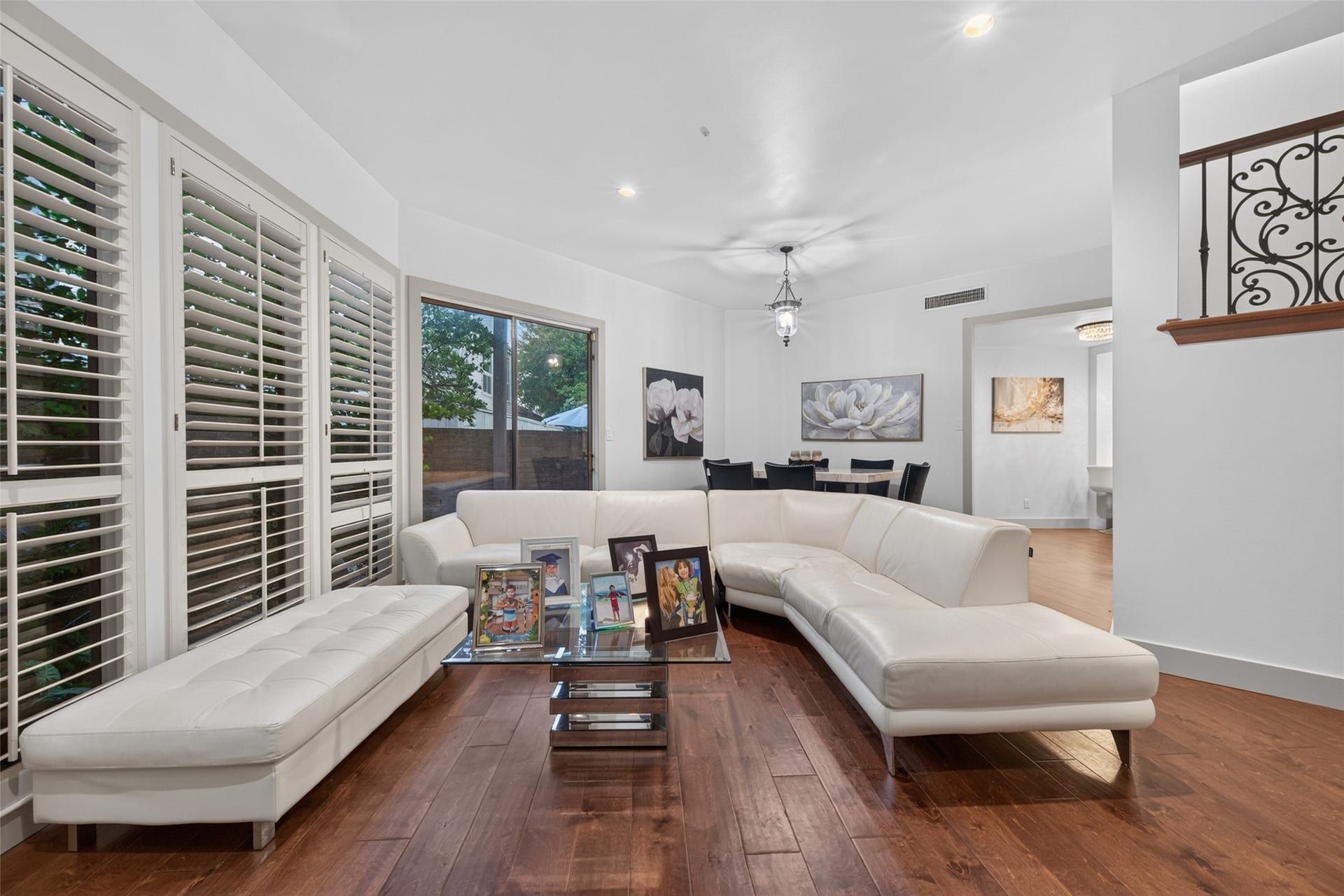 This inviting living area features a modern white sectional sofa, engineer wood floors, and abundant natural light from large windows with shutters. The open layout includes a dining space with stylish lighting and elegant artwork.
