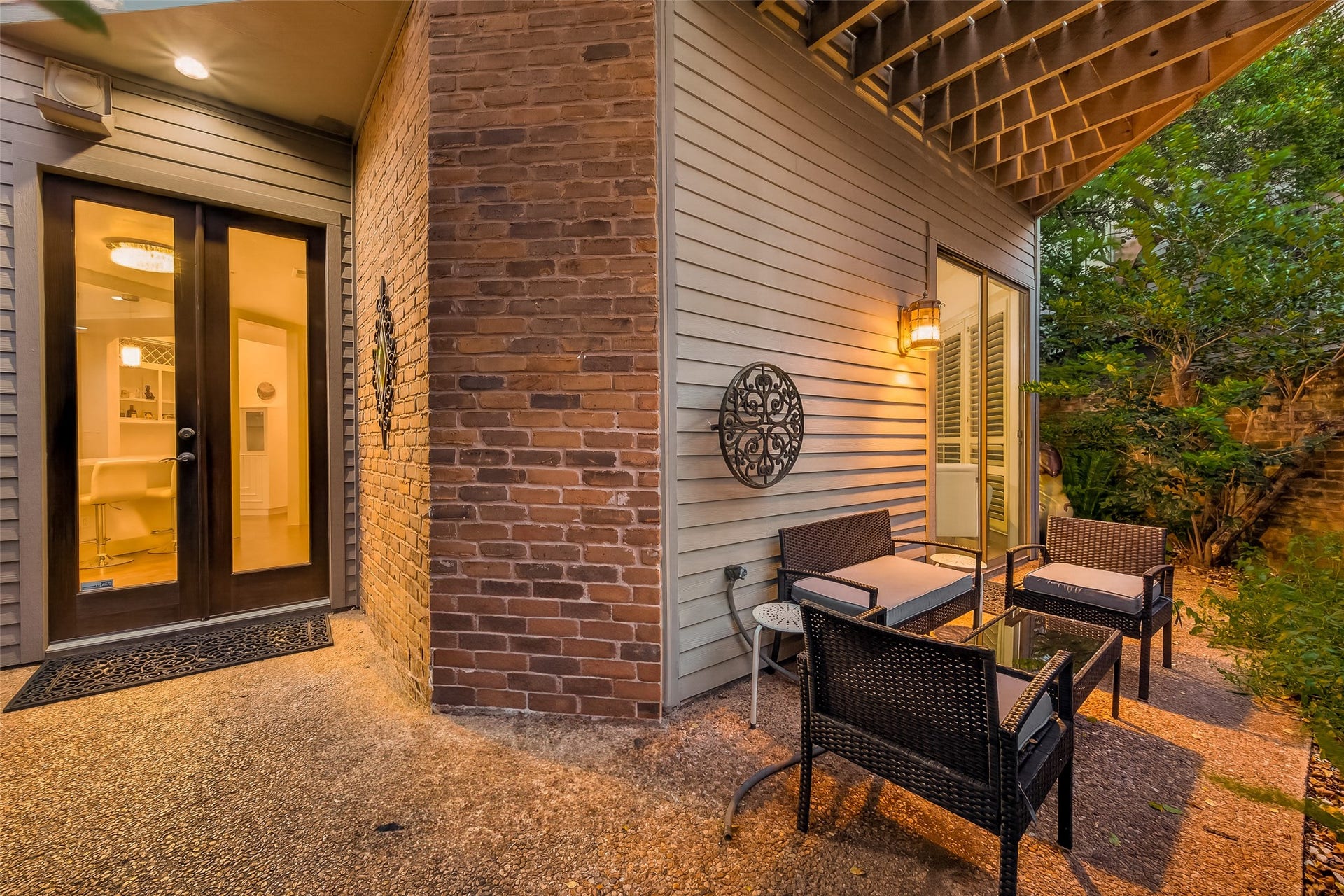 This photo showcases a cozy outdoor patio area with comfortable seating and a glass-top table, set against a backdrop of brick and siding walls. The space is warm and inviting, enhanced by ambient lighting and surrounded by lush greenery, perfect for relaxation or entertaining.
