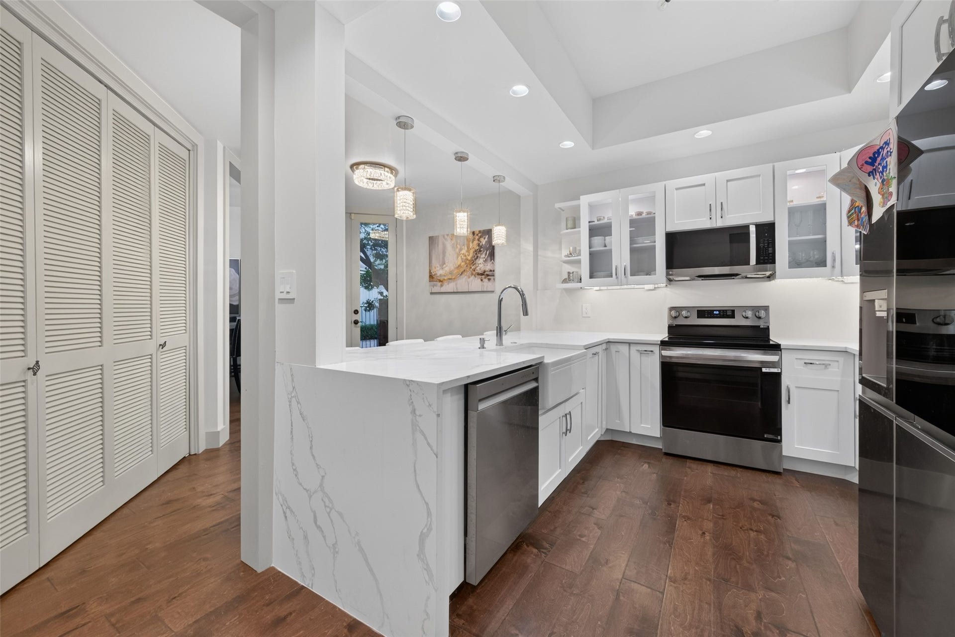 This modern kitchen features sleek white cabinetry, stainless steel appliances, and a stylish marble countertop. The space is brightened by recessed lighting and pendant lights, with warm hardwood floors adding a cozy touch.
