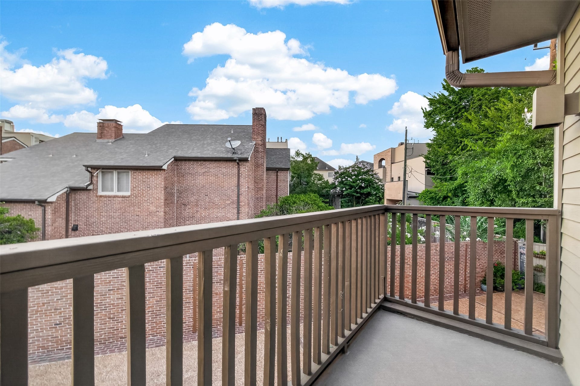 A small balcony with a wooden railing overlooks a courtyard surrounded by brick buildings and greenery, providing a cozy outdoor space.