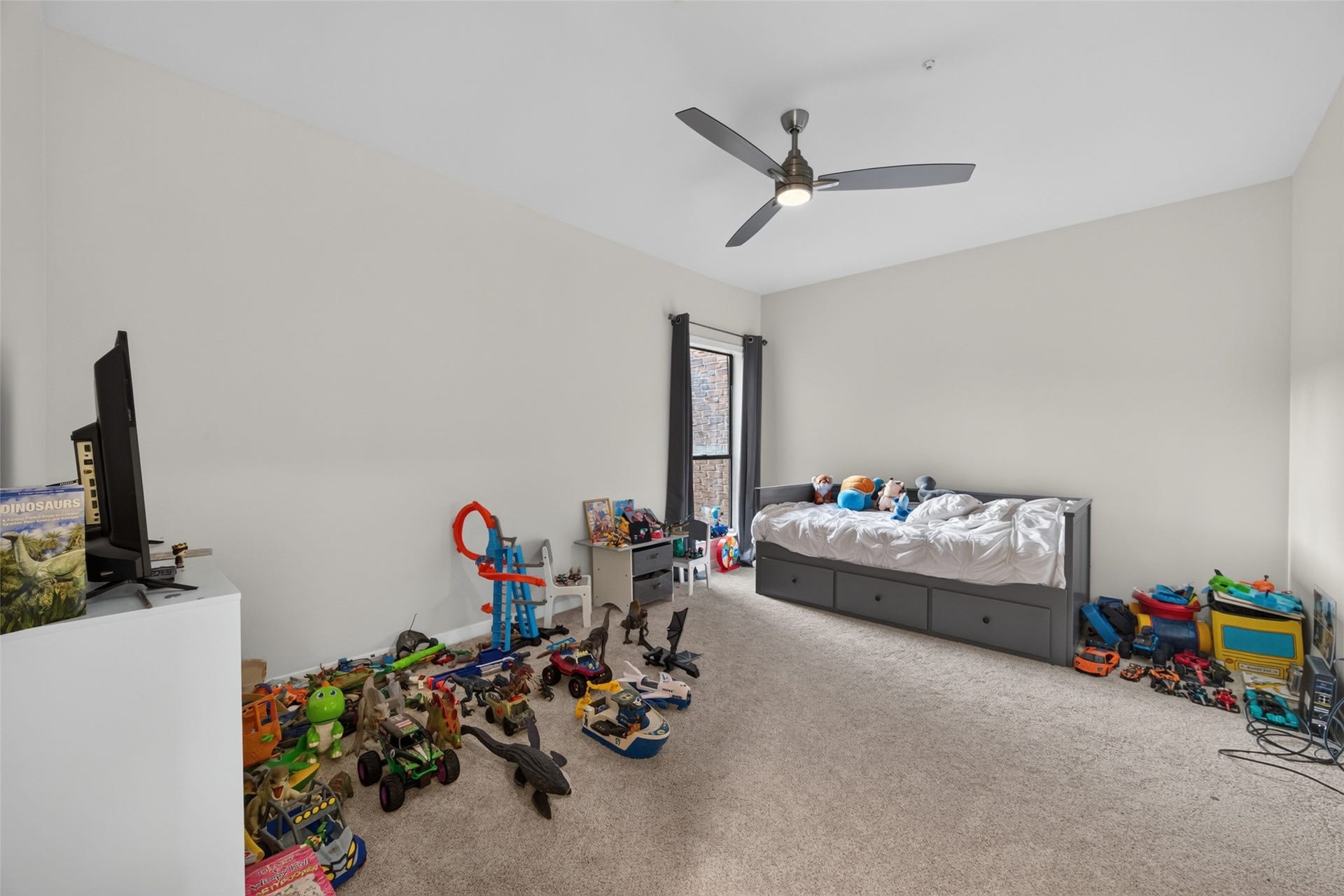 This spacious bedroom #2 features neutral walls, carpeted floors, and a ceiling fan. It includes a bed with storage drawers underneath and ample space for toys, making it ideal for a child's room. Natural light enters through a large window with dark curtains.