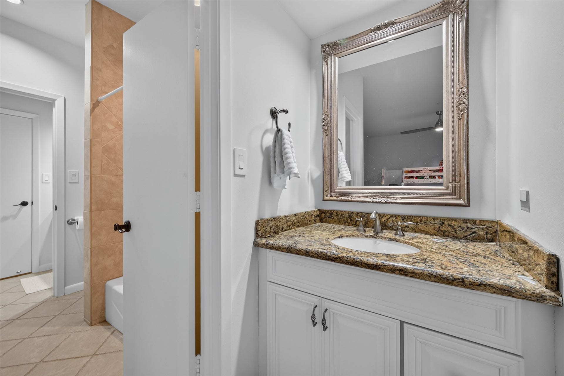This photo shows a bathroom with a granite countertop and a stylish framed mirror above the sink. On the left, there's a partial view of a shower with tan tiles and a white bathtub. The space features neutral tones and modern fixtures.