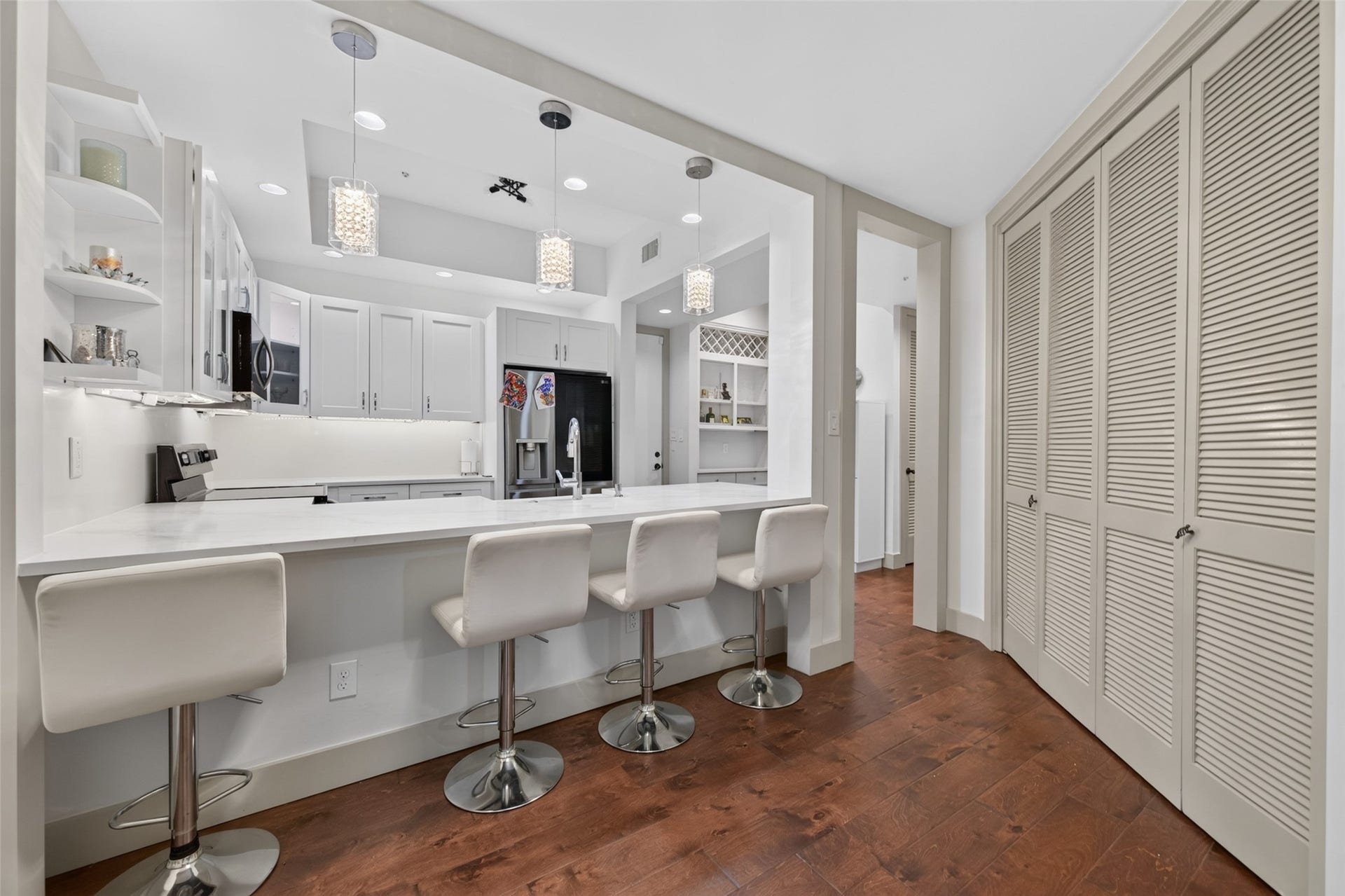 This photo showcases a modern kitchen with white cabinetry, a sleek countertop, and bar seating for four. It features pendant lighting, stainless steel appliances, and ample storage space, creating a stylish and functional cooking area.