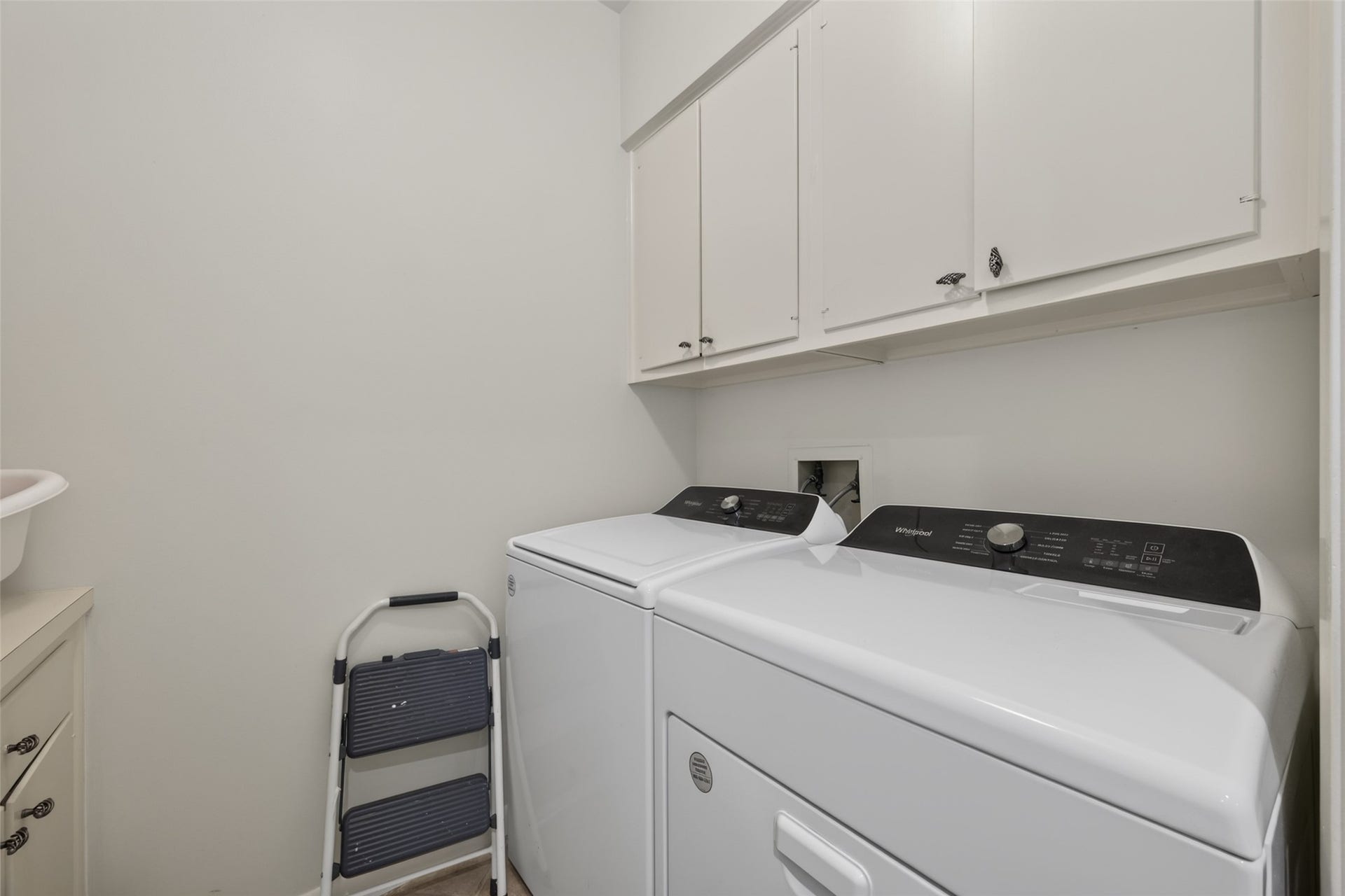 This photo shows a bright, functional laundry room featuring a washer and dryer, upper storage cabinets, and a small step ladder for easy access.