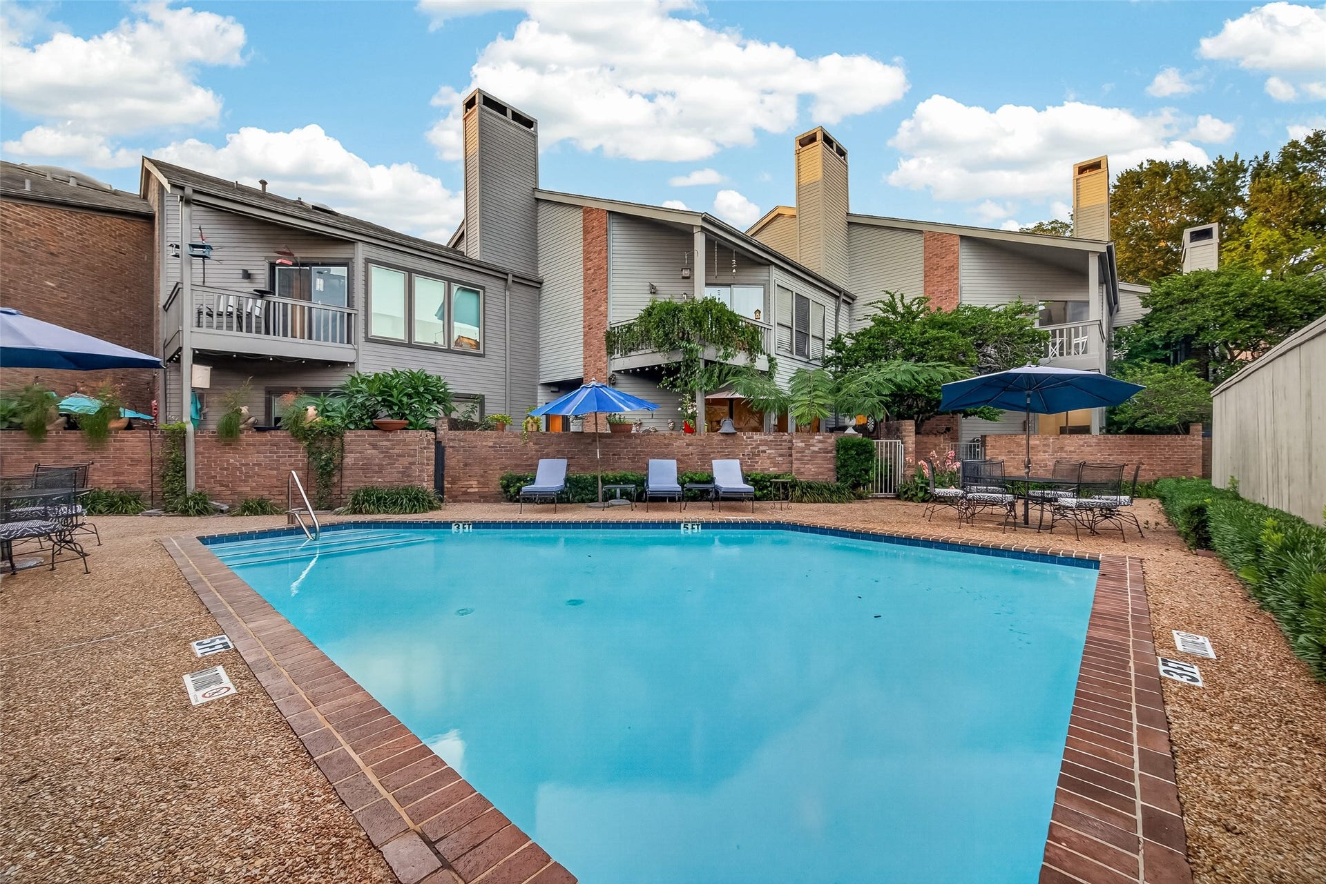 This photo features a serene outdoor area with a sparkling swimming pool surrounded by a stone patio. The property includes multi-level buildings with private balconies and lush greenery. Ideal for relaxation and entertaining.