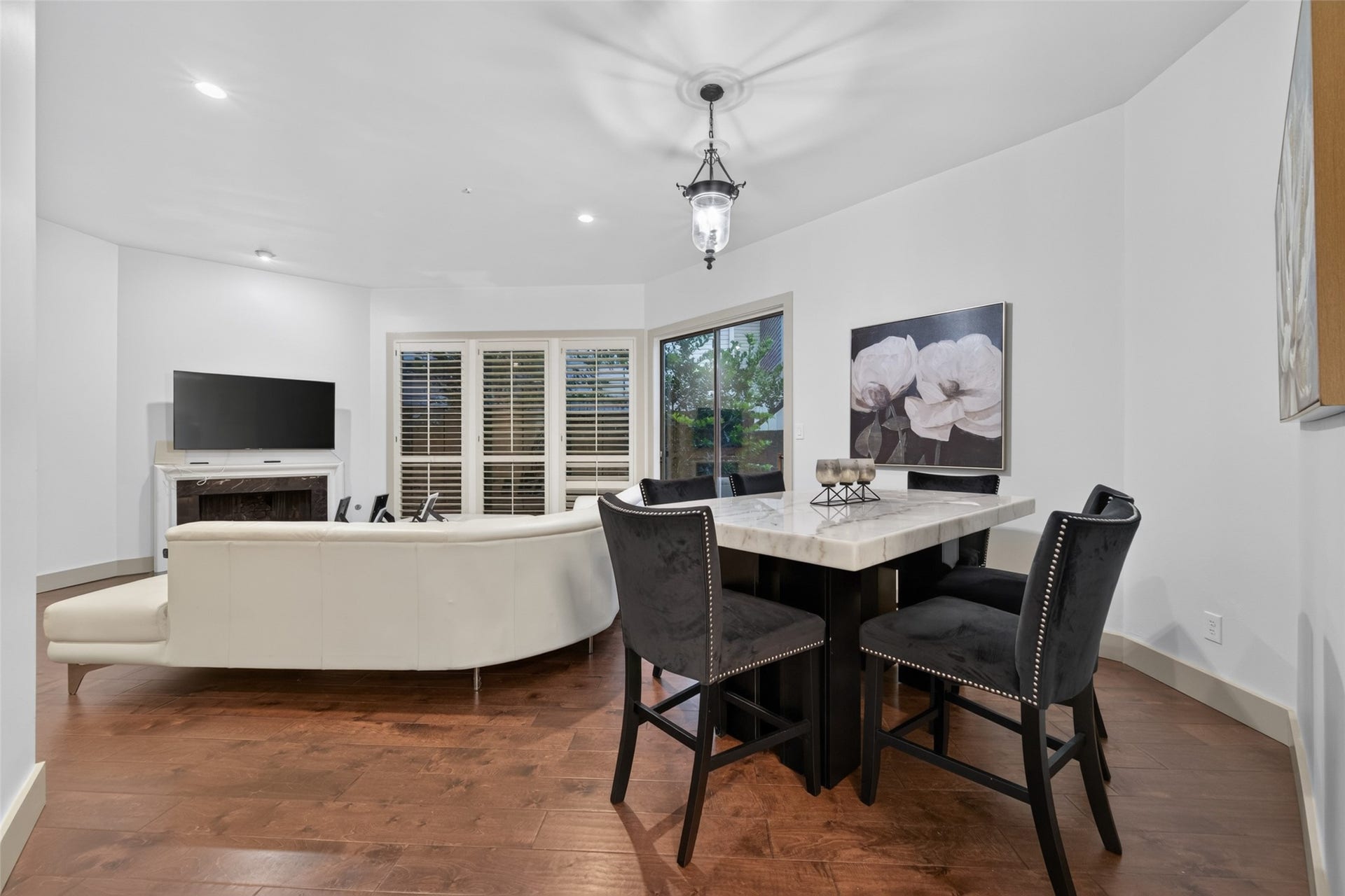 This inviting living and dining area features engineer wood  floors, a cozy white sofa, and a mounted TV. The dining space boasts a stylish table with four chairs, while large windows with shutters and a sliding glass door provide ample natural light.