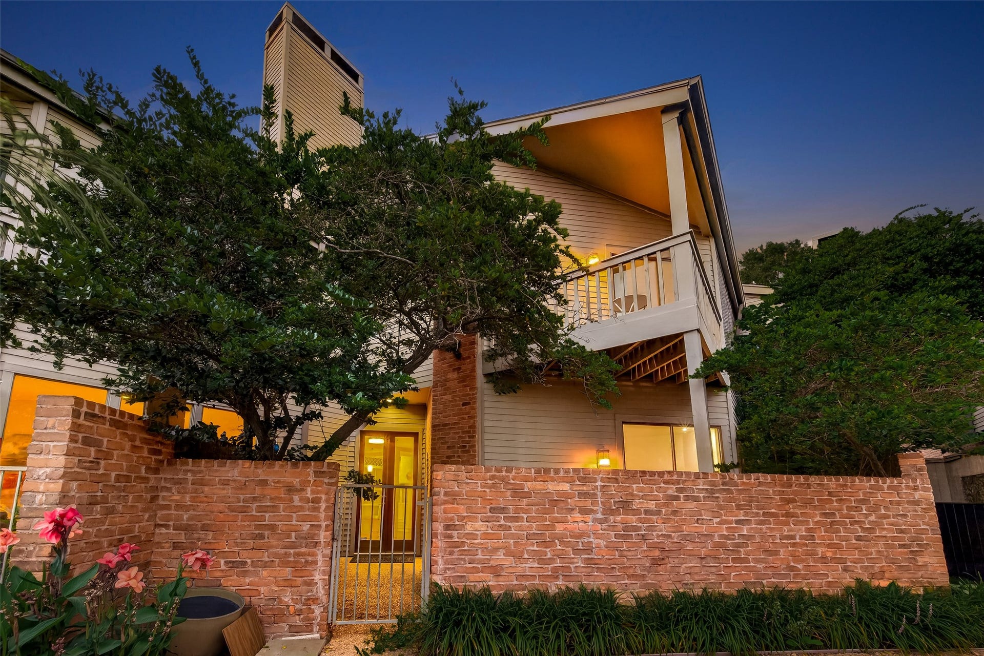 This inviting home features a charming brick exterior with a private patio area. The warm lighting complements the lush greenery and the cozy porch, creating an appealing and welcoming atmosphere.