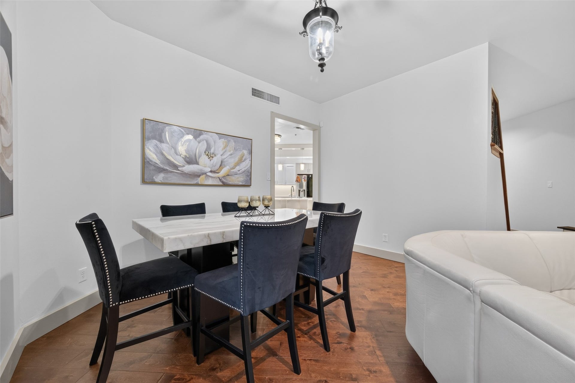 This inviting dining area features a modern white table with six navy chairs, accented by elegant wall art and a central ceiling light. The space flows seamlessly into the adjacent room, offering a glimpse of a stylish kitchen.