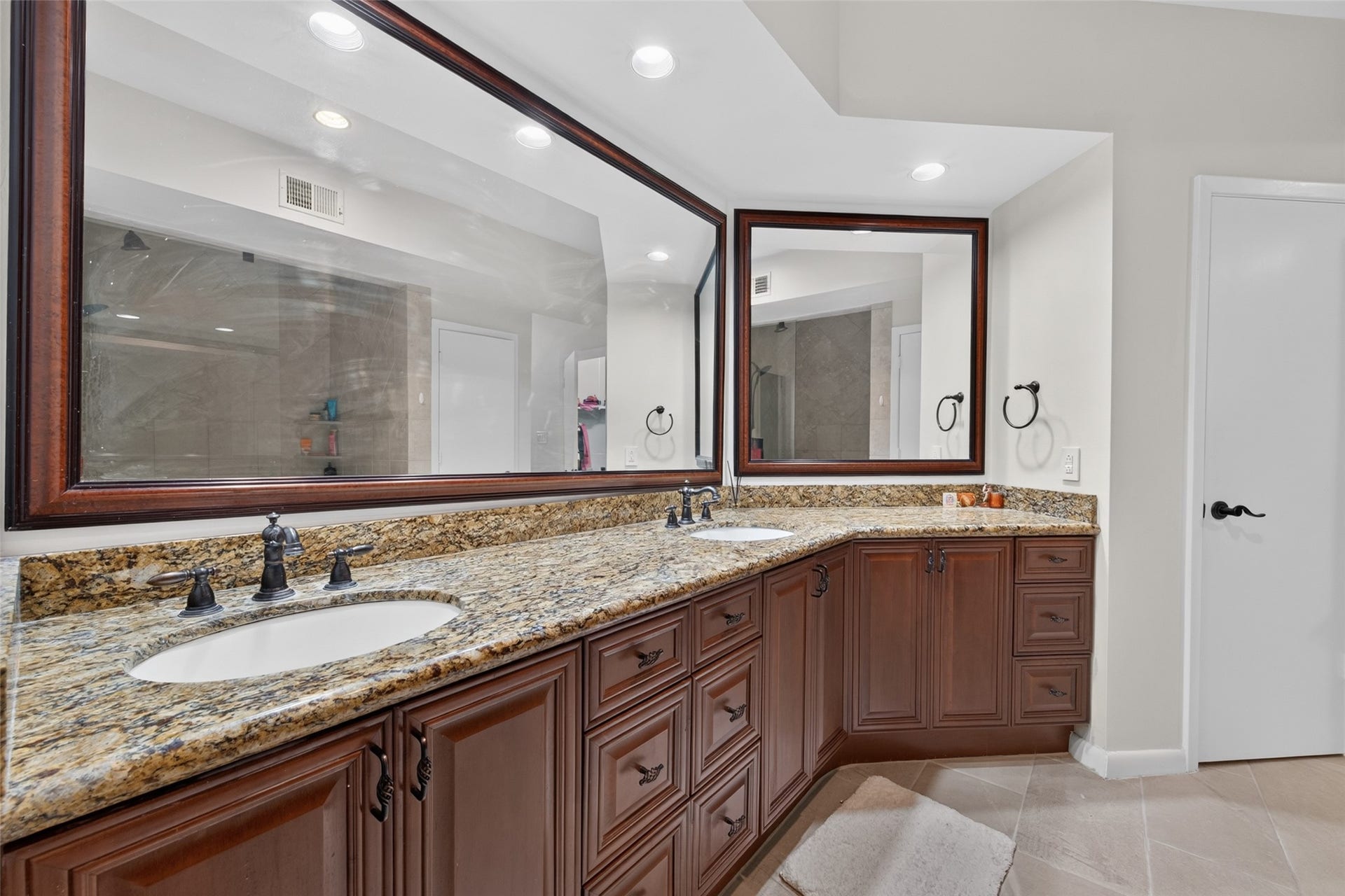This photo showcases a spacious bathroom with a dual-sink vanity featuring granite countertops and rich wooden cabinetry. Large mirrors and recessed lighting enhance the space, offering a luxurious and functional design.