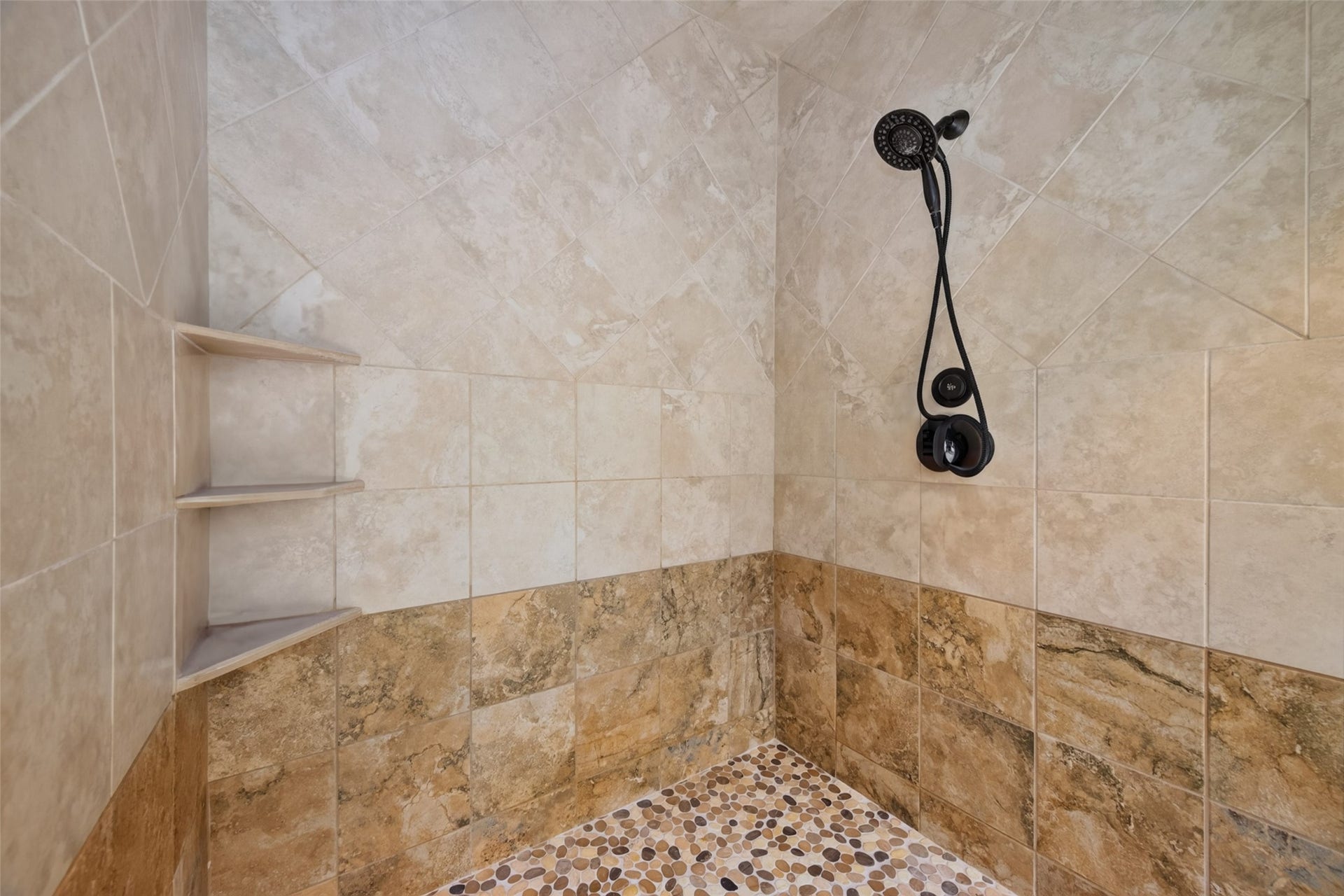 This photo shows a stylish shower with beige and brown tiles, featuring a modern rainfall showerhead and built-in corner shelves. The pebble stone floor adds a natural touch to the space.