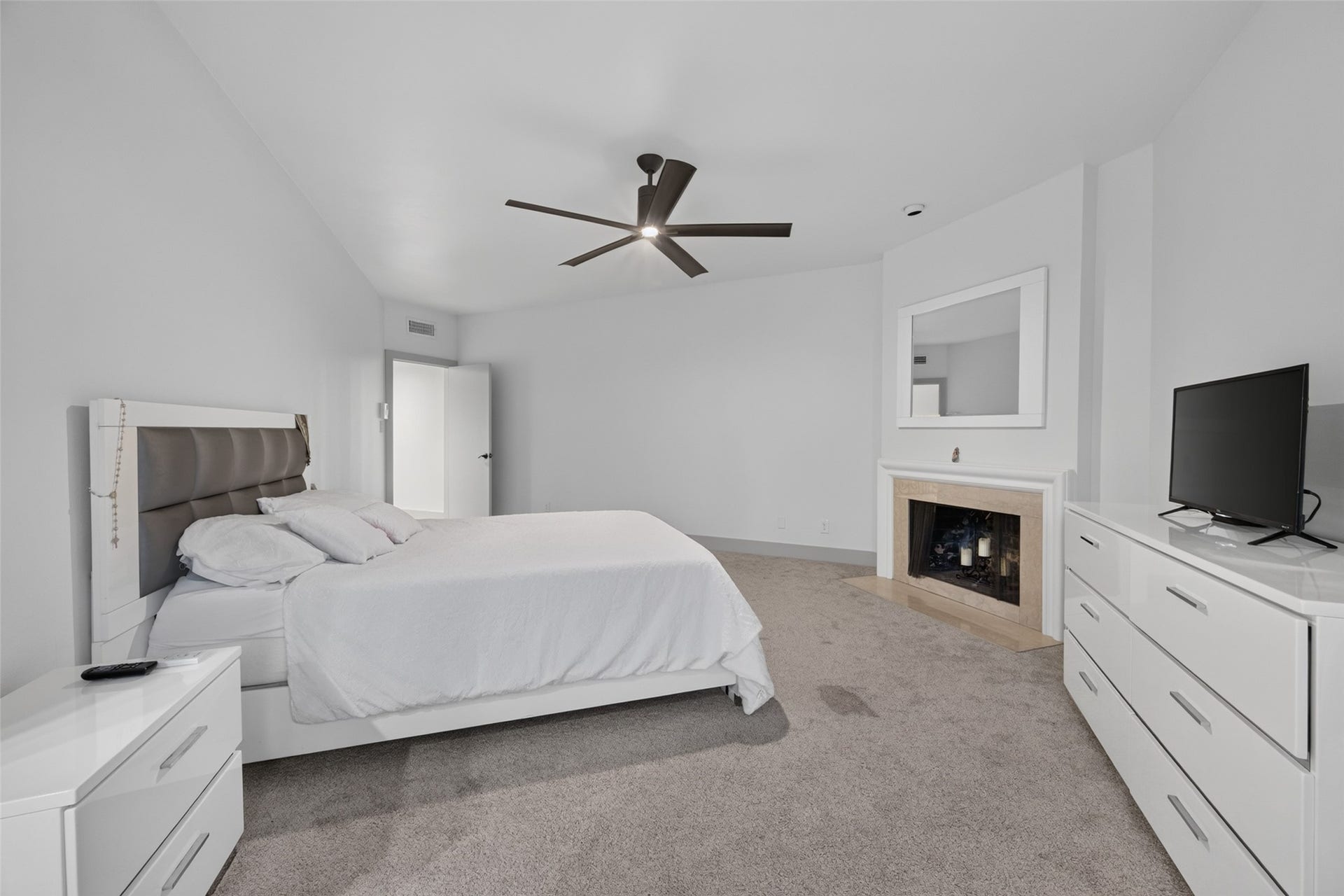 This spacious bedroom features a modern ceiling fan, a cozy fireplace, and neutral carpeting. It includes a large bed, ample dresser space, and a wall-mounted TV, creating a comfortable and inviting atmosphere.