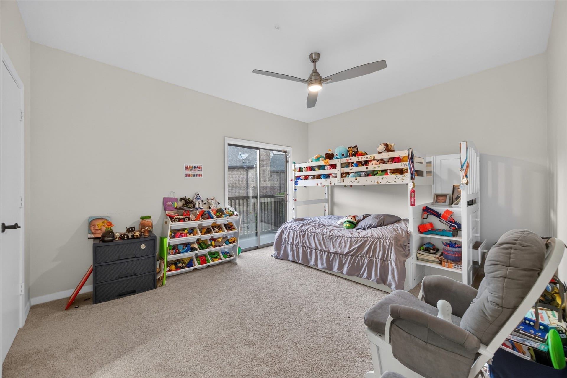 This spacious, carpeted bedroom #3 features a modern ceiling fan, sliding glass door to a balcony, and is ideal for a child's room with built-in bunk beds, toy storage, and a cozy rocking chair.