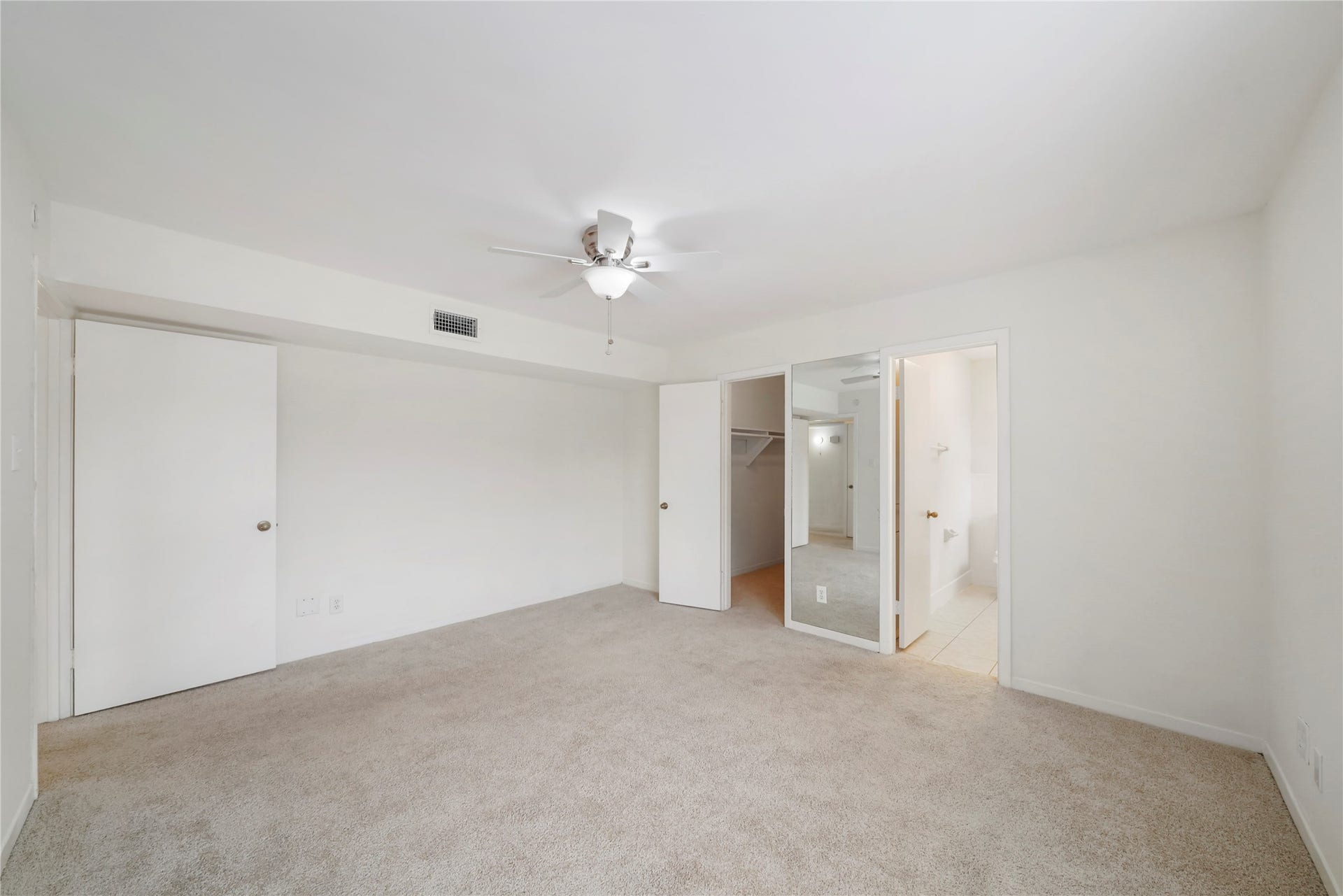 Primary bedroom showing built-in closet storage, mirrored sliding doors, direct bathroom access, a ceiling fan, and soft carpeting throughout.