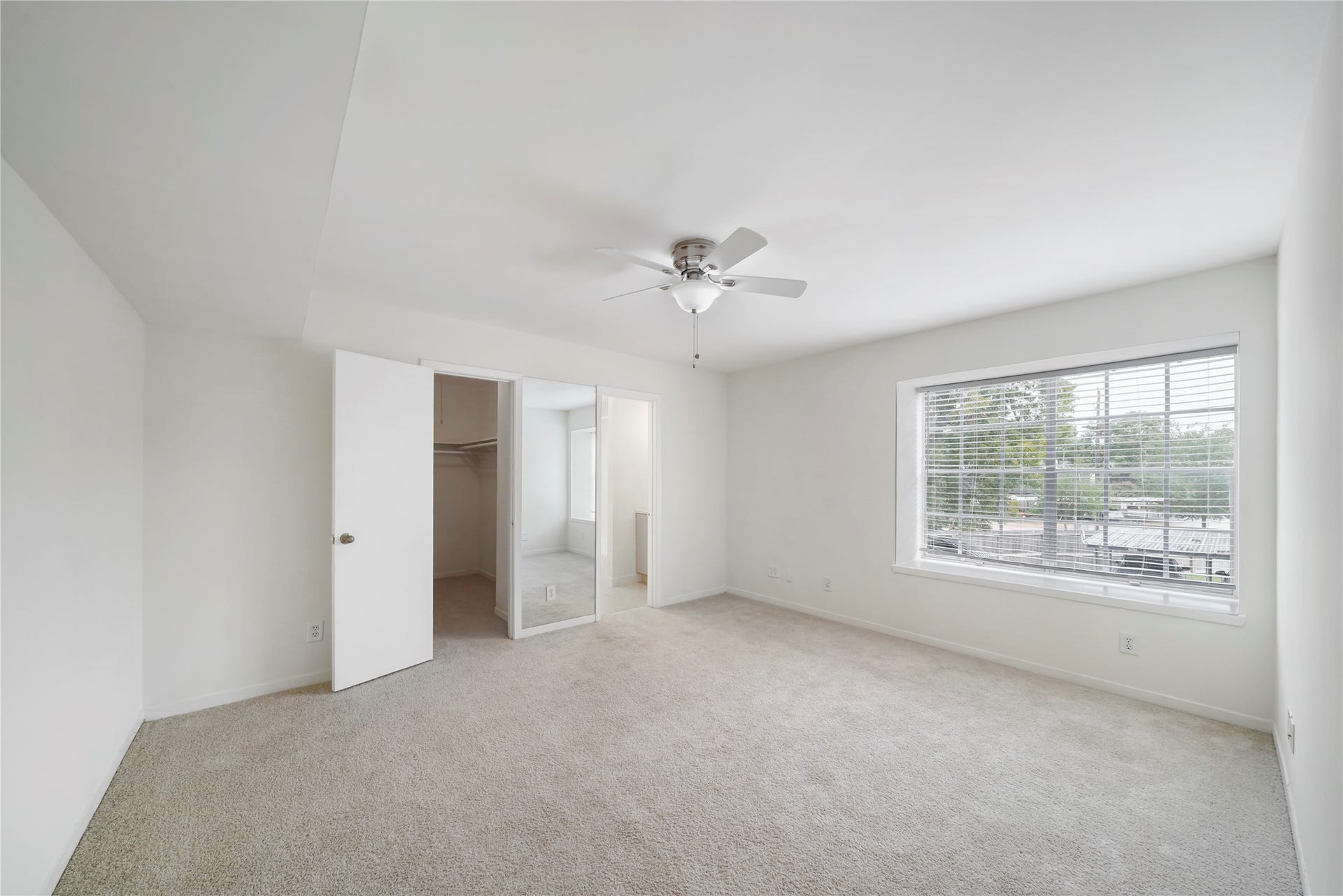 Generous bedroom suite equipped with a ceiling fan, broad window with blinds, walk-in closet, mirrored secondary door, and plush carpet.