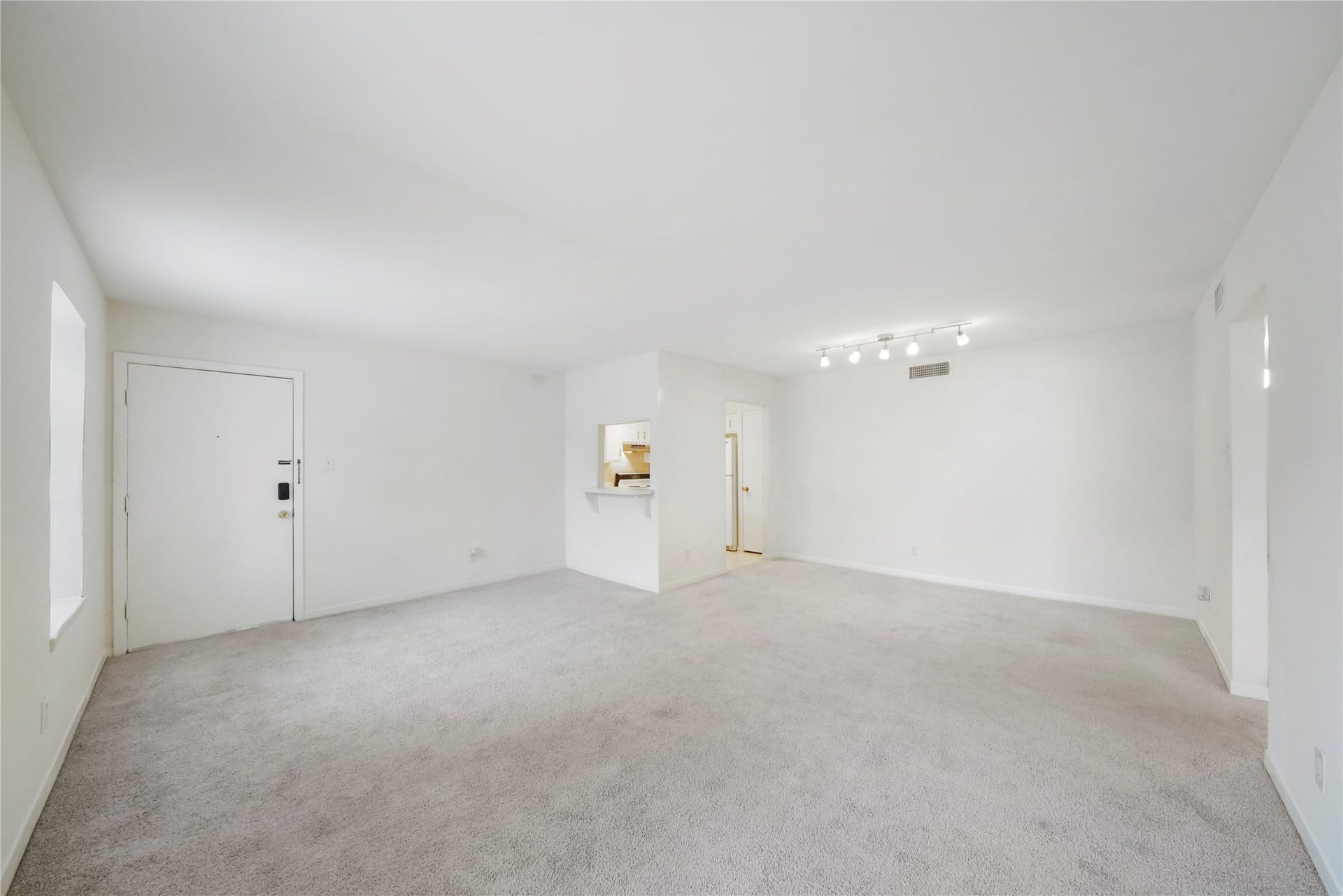 Open-concept living room with neutral carpet, bright natural light from adjacent windows, entry access, and a sightline into the kitchen area.